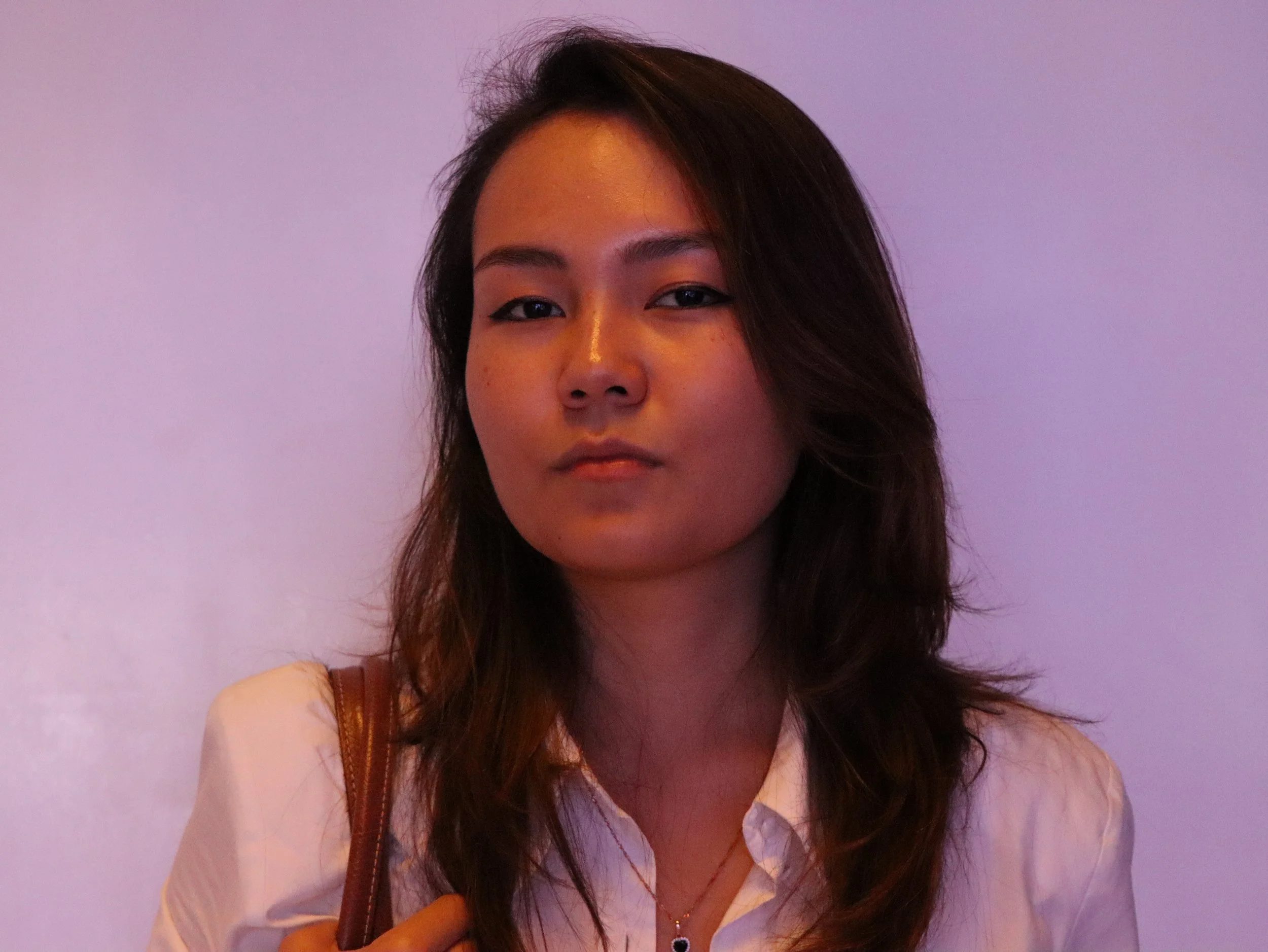 A young woman with long dark hair and a serious expression, wearing a white shirt and a brown shoulder bag, standing against a plain lilac background.