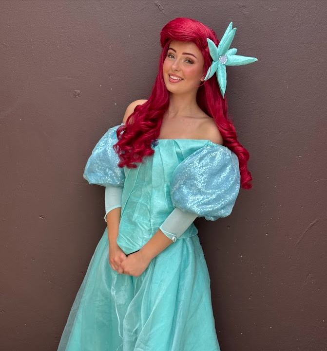 Young woman dressed as a princess with red hair, wearing a blue gown with puffy sleeves and a tiara, standing outdoors in front of lush purple and green foliage.