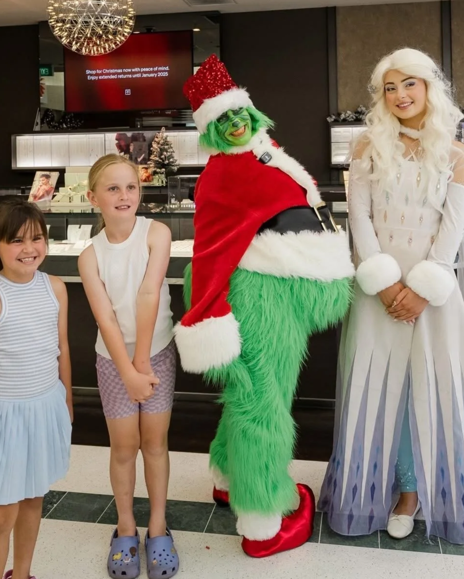 Three children and two women in festive costumes posing for a photo in a store. The women are dressed in holiday-themed outfits, one as a Christmas elf and the other as a winter fairy. The children are smiling and standing with them.