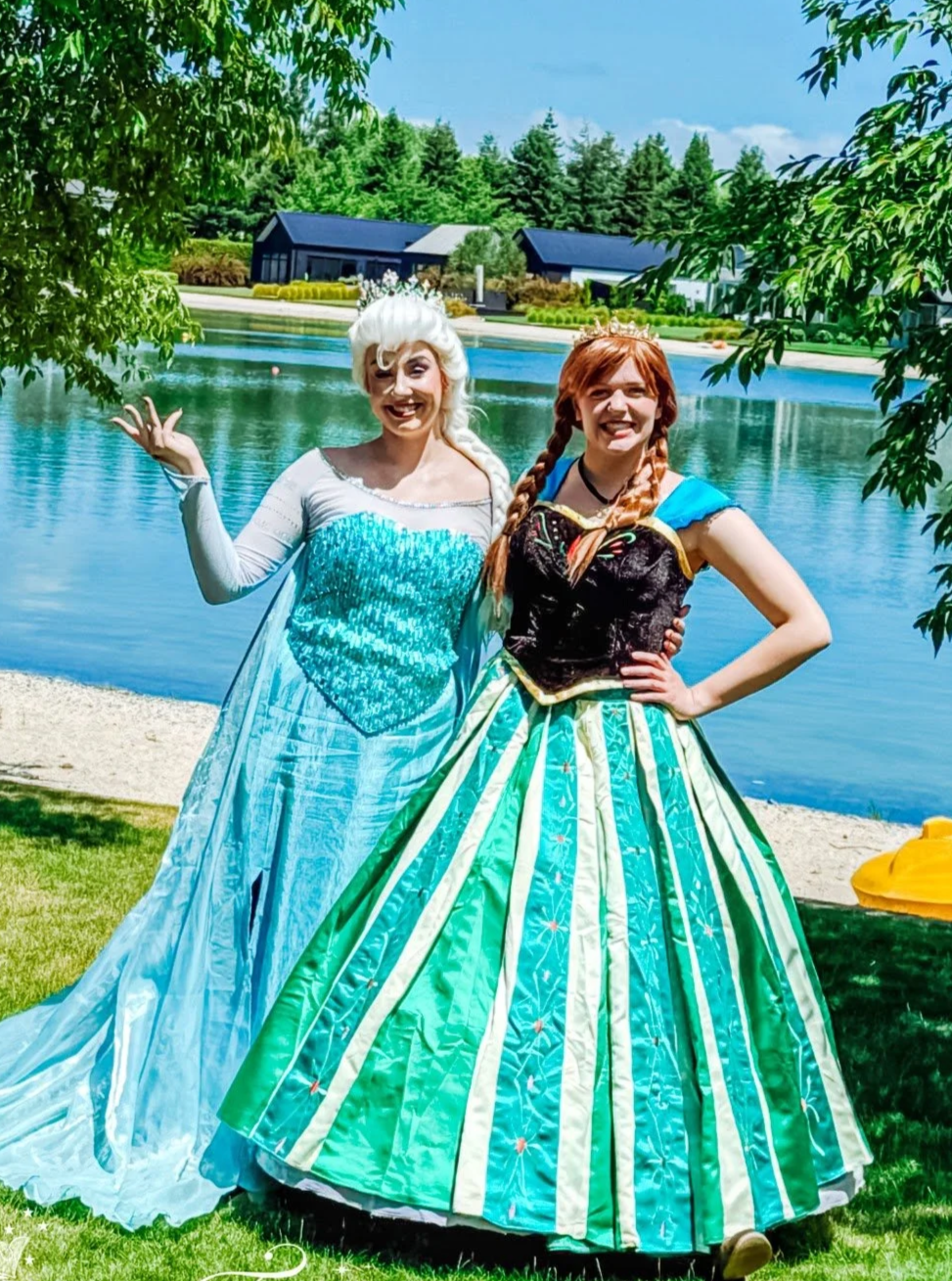 Two people in costumes at an indoor event, with one dressed in a purple cape and crown, and the other in a white dress with long white hair. The background shows people and vendor booths. Hire Elsa and Anna Rangiora)