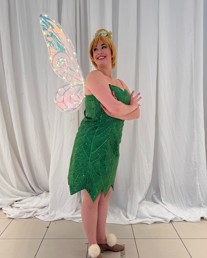Woman dressed as Tinker Bell, wearing a green dress and fairy wings, smiling and posing with her hands on her hips.