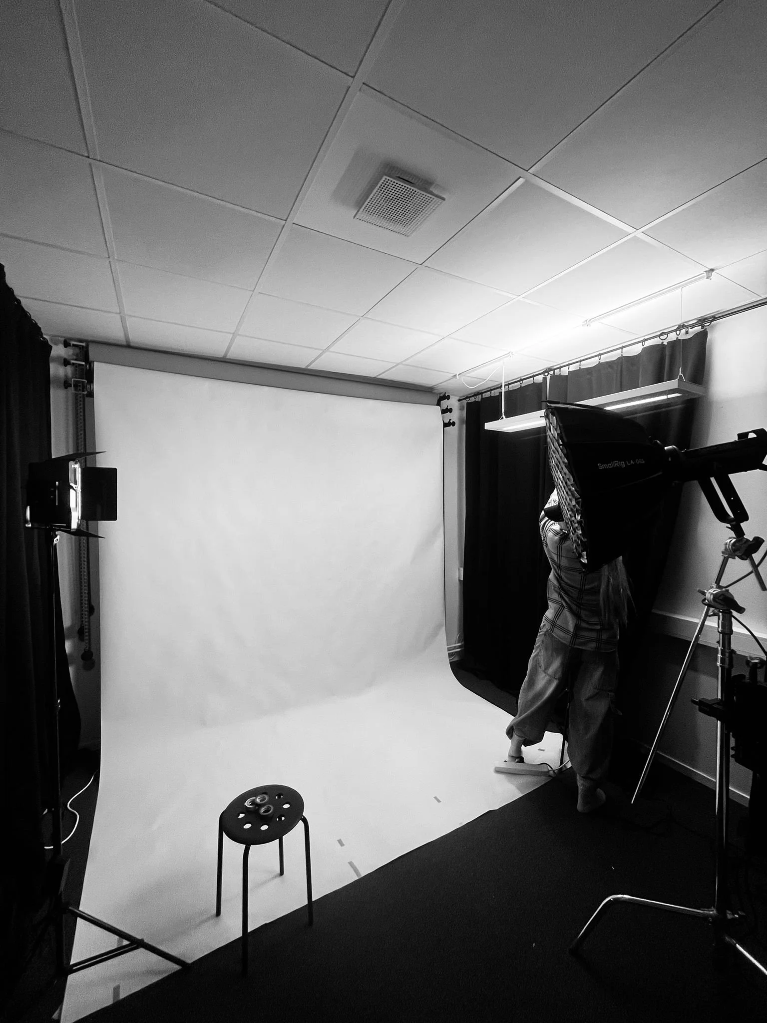 Fotostudio med vit bakgrund, fotograf som justerar belysning, studioutrustning, svart och vitt tyg som gardiner, stol och mörkt golv.