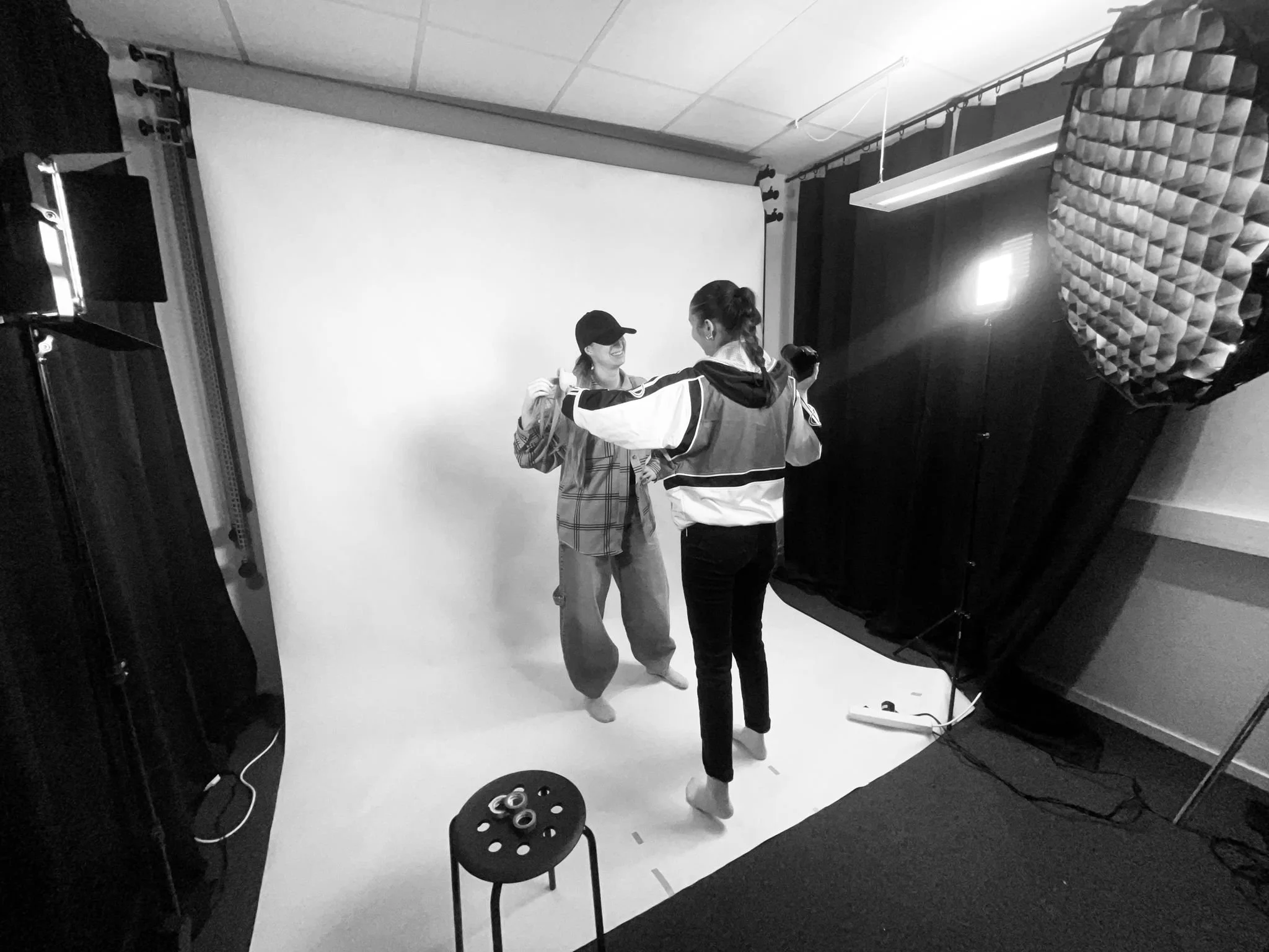 Två kvinnor står på en fotostudio med ett vitt bakgrundsblad, en av dem är med fällbar jacka och den andra med keps, de håller händerna mot varandra.