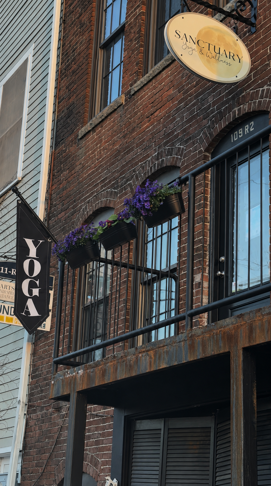 Exterior of a brick building with signs indicating a yoga and wellness studio and a balcony with purple flowers in hanging planters.