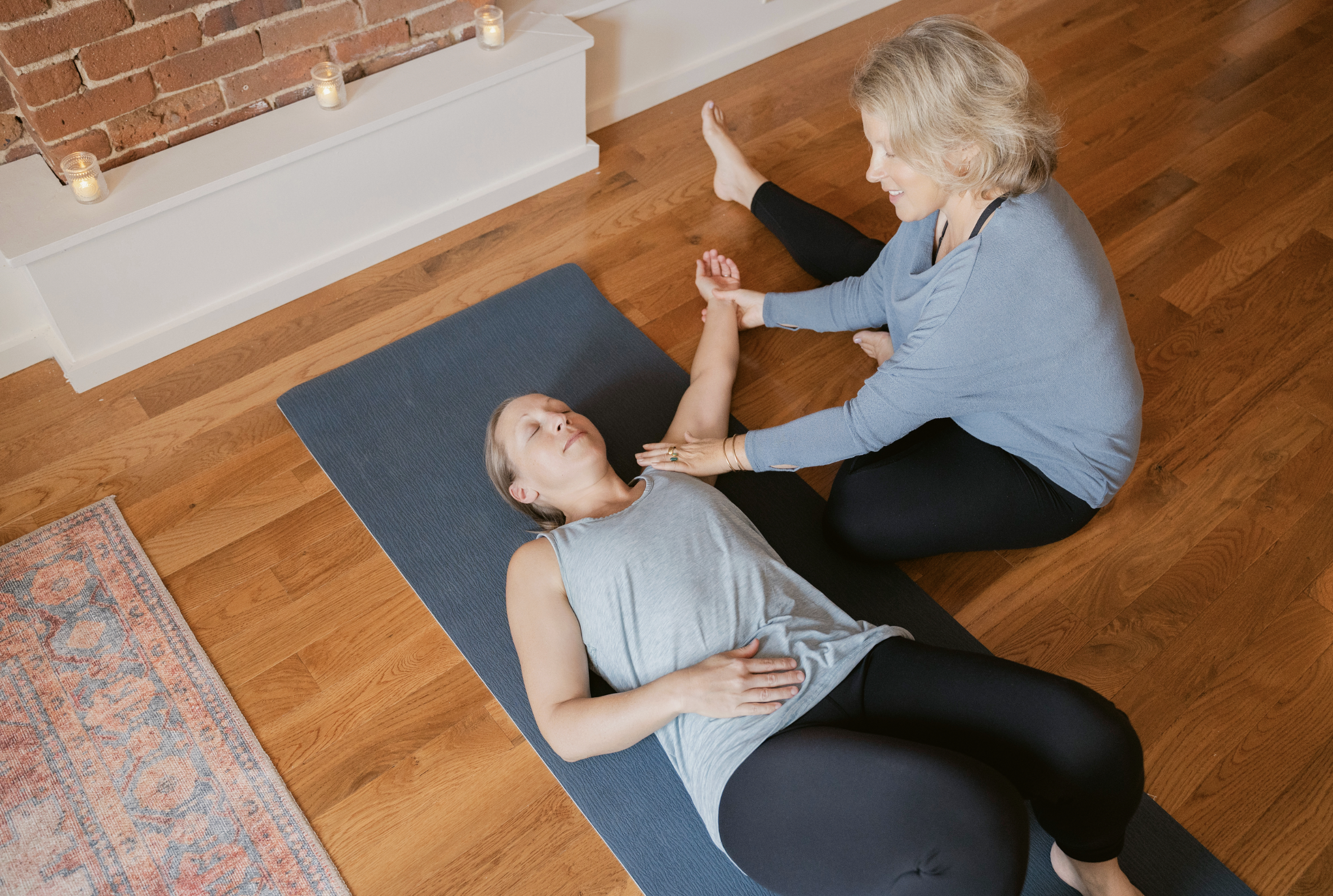 A woman practicing yoga or meditation with a child on a yoga mat in a cozy room with hardwood floors, a brick fireplace, and candles.