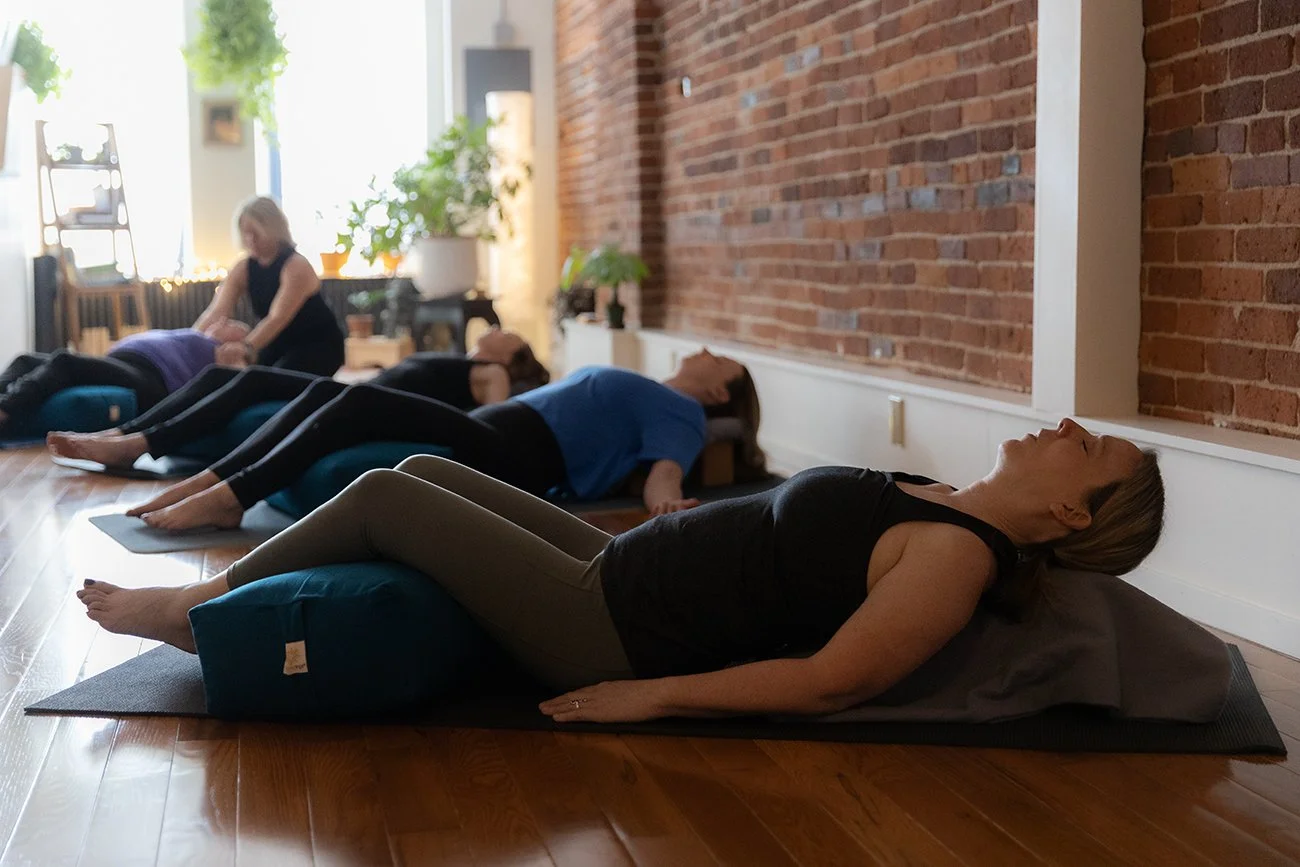 People practicing yoga in a studio with a brick wall, lying on mats with pillows, with a woman guiding the session.