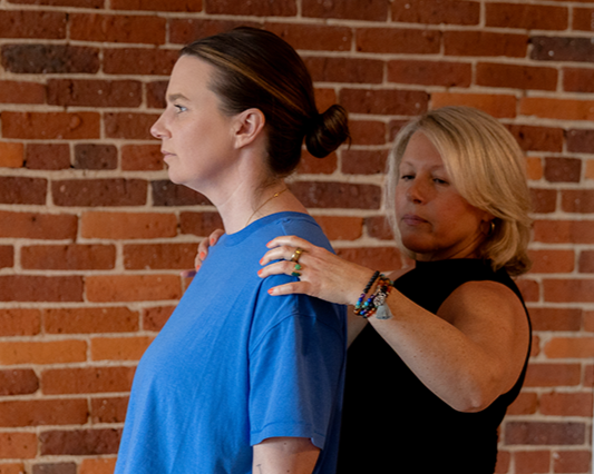 A woman receives a shoulder massage from another woman against a brick wall background.
