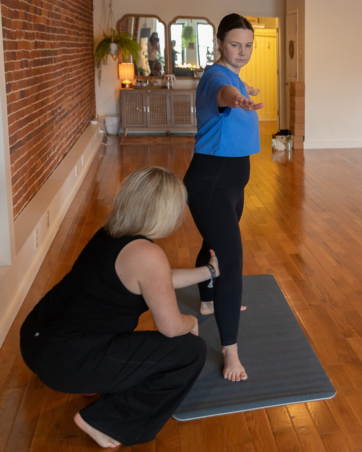 A woman with dark hair in a blue shirt practices yoga on a gray mat while an instructor helps her with her pose in a cozy living room with wooden floors and a brick accent wall.