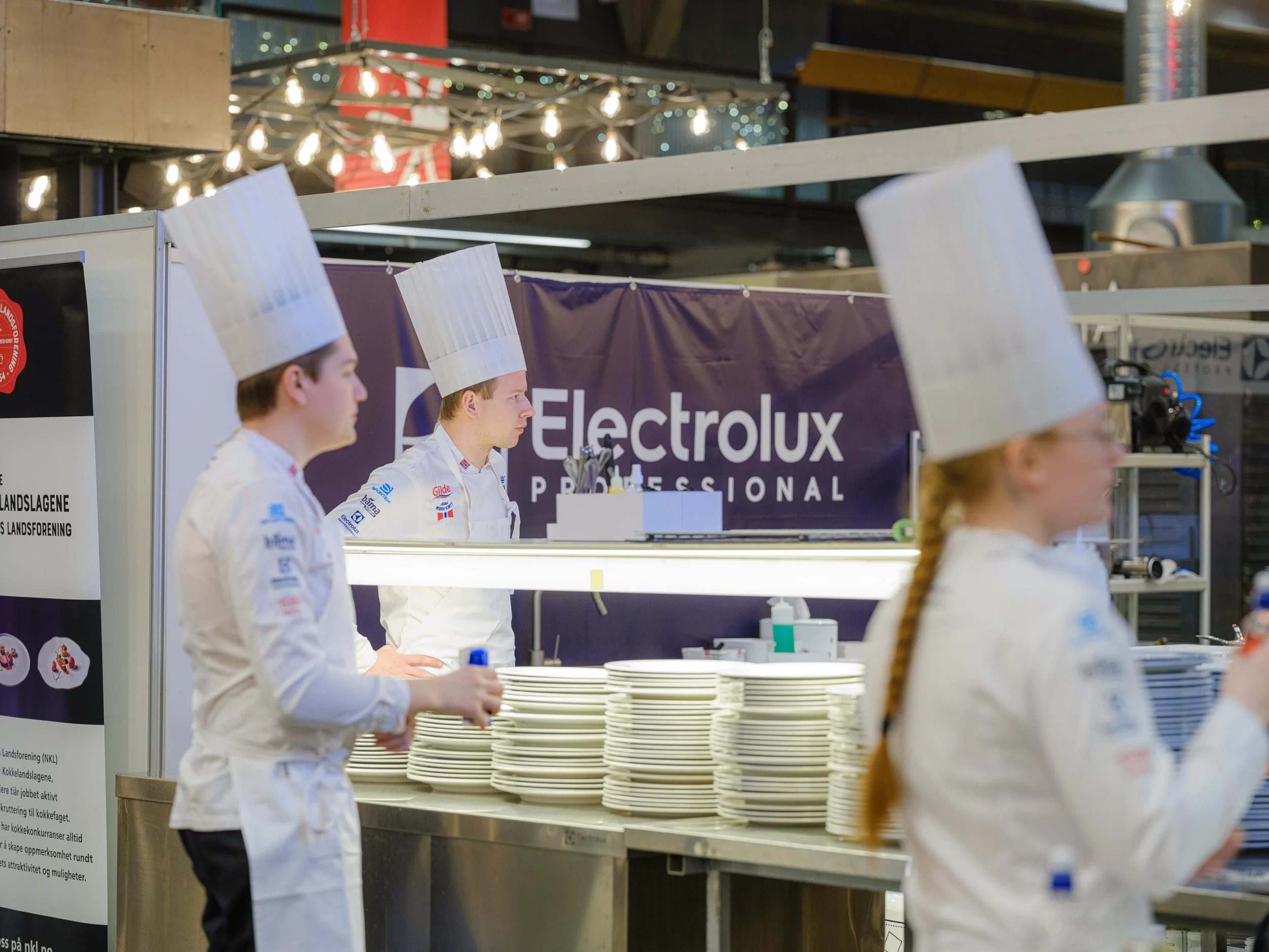 Kokker i uniformer med høye kokkeluer står ved et kjøkkenområde med Electrolux-logo i bakgrunnen. Det er stabler med tallerkener foran dem.