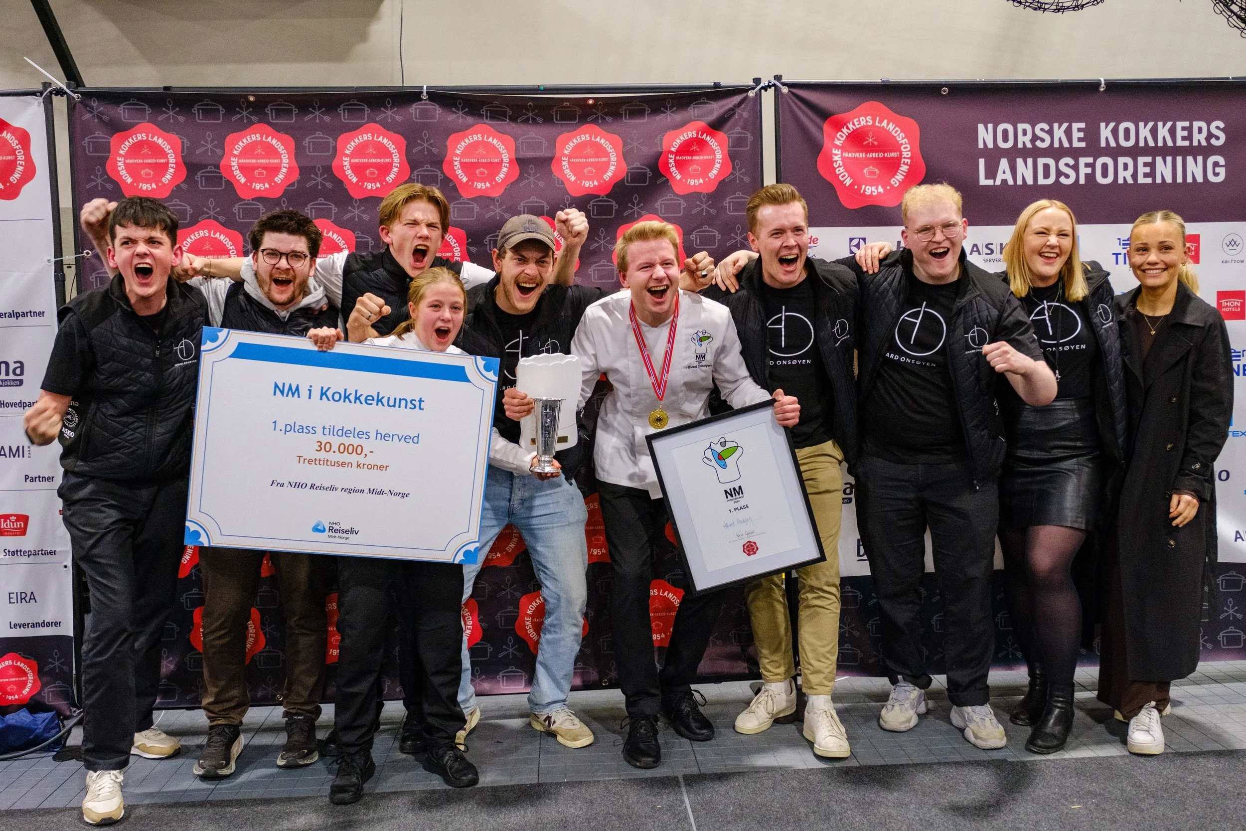 Håvard Onsøyen og hans team feirer seier i NM i kokkekunst, holder et stort sjekk og trofé. Bakgrunnen viser bannere med logoer fra Norske Kokkers Landsforening.