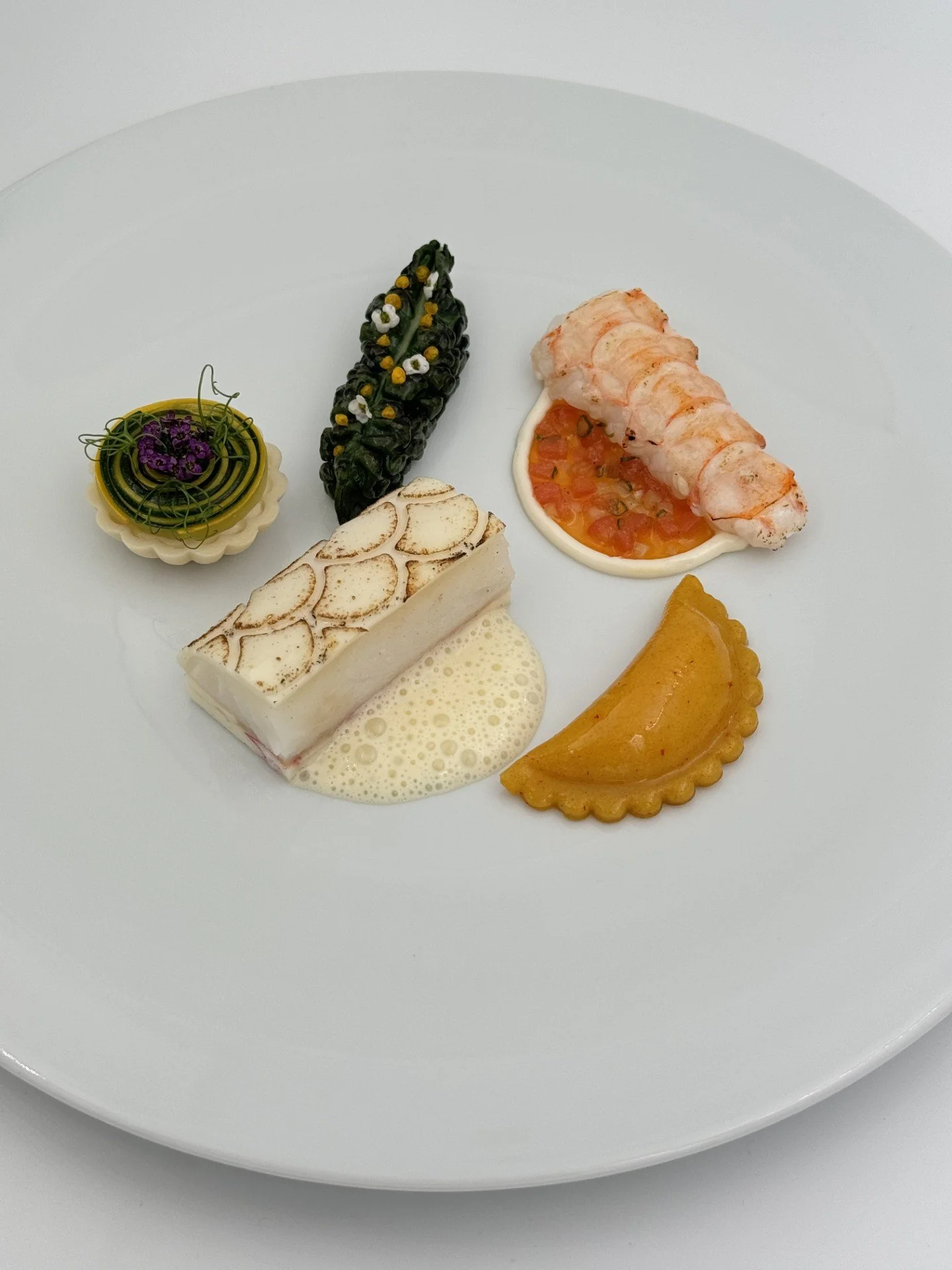 Gourmetrett med ulike små porsjoner, inkludert en bit hvit fisk på skumsaus, en grillet sjøkreps på tomat- og sausbase, en fylt pastabit, en spiralisert grønn grønnsakstert, og en dekorert grønn bladerull.