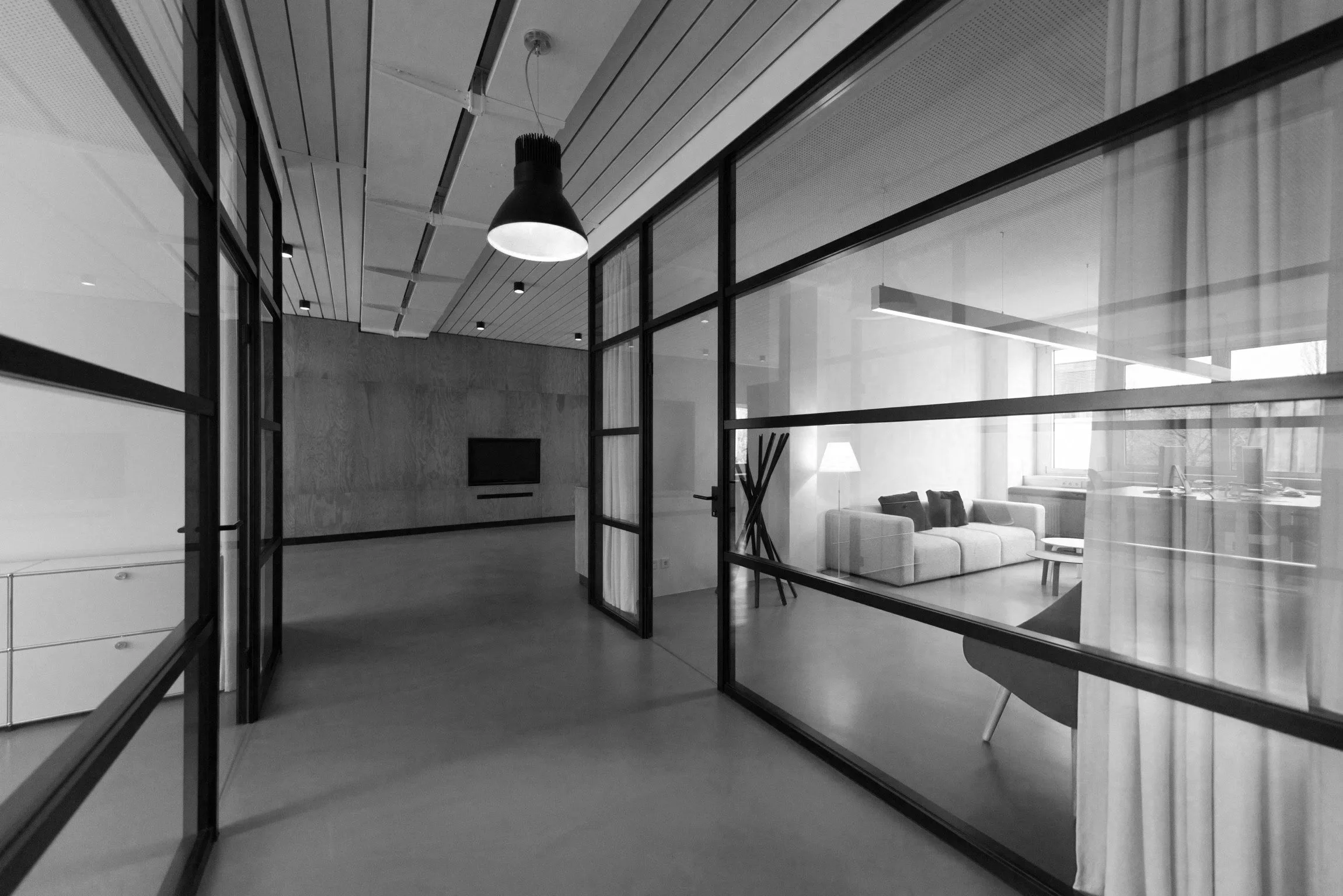 Interior black and white photo of modern office space with glass partitions, a seating area with a sofa and a lamp, ceiling lights, and large windows.