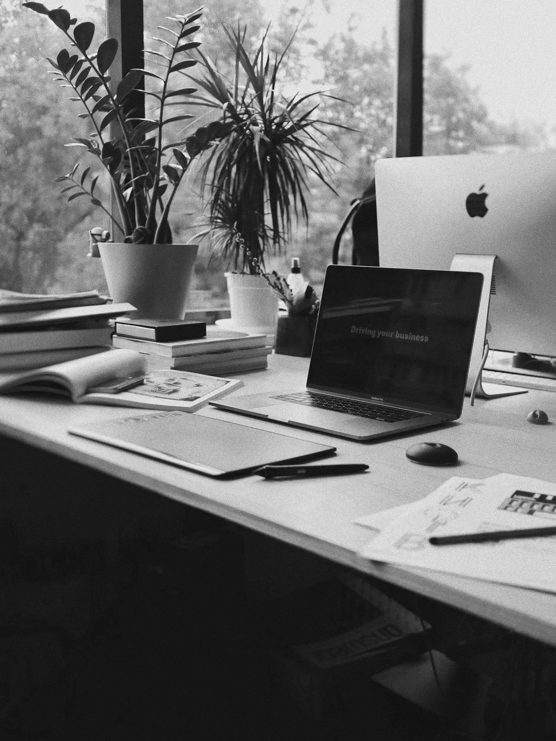 A cluttered workspace with a desk, plants, books, a laptop, a tablet showing 'Driving your business' and various papers and office supplies.