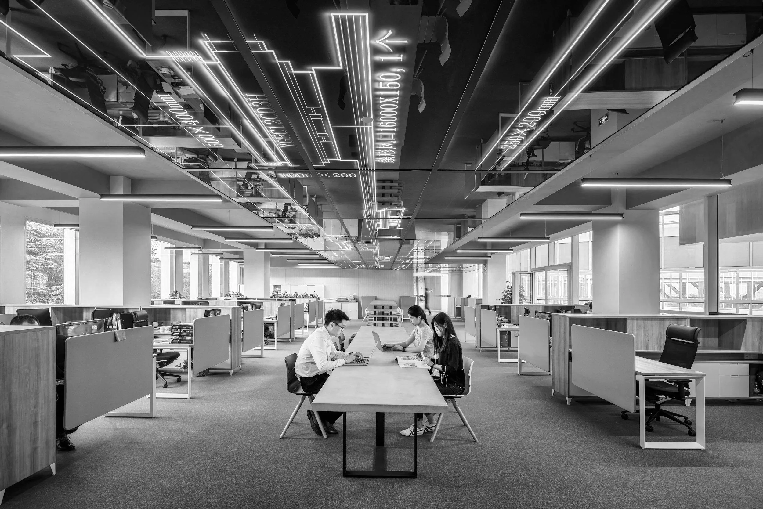 Inside a modern office with large windows and ceiling lights, three young professionals sit at a table working on laptops, with glass partitions and office furniture visible.