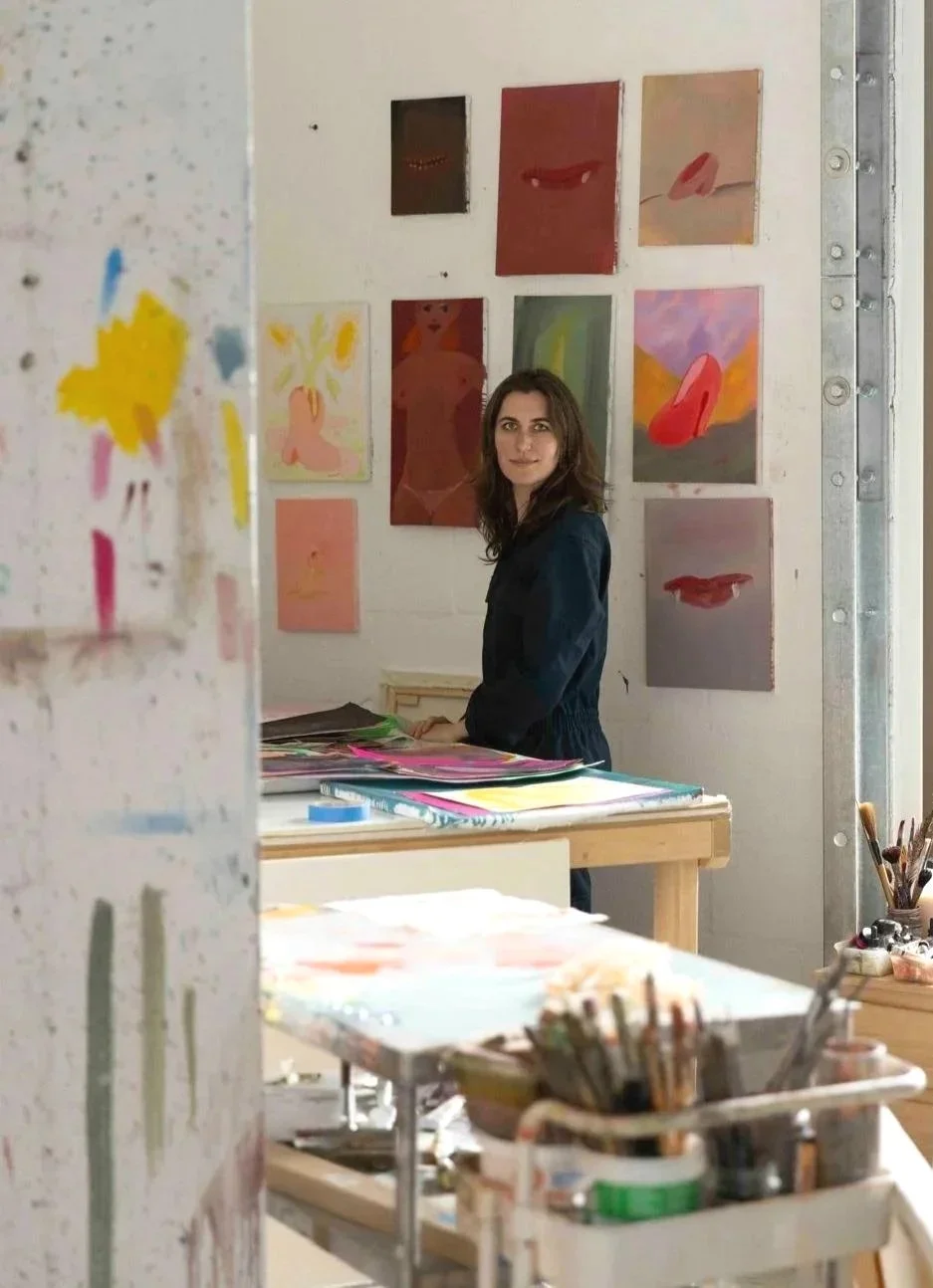 A woman with brown hair wearing a black jacket, standing in an art studio with paintings on the wall behind her. Art supplies, brushes, and stacks of paper are visible on the tables in the foreground.