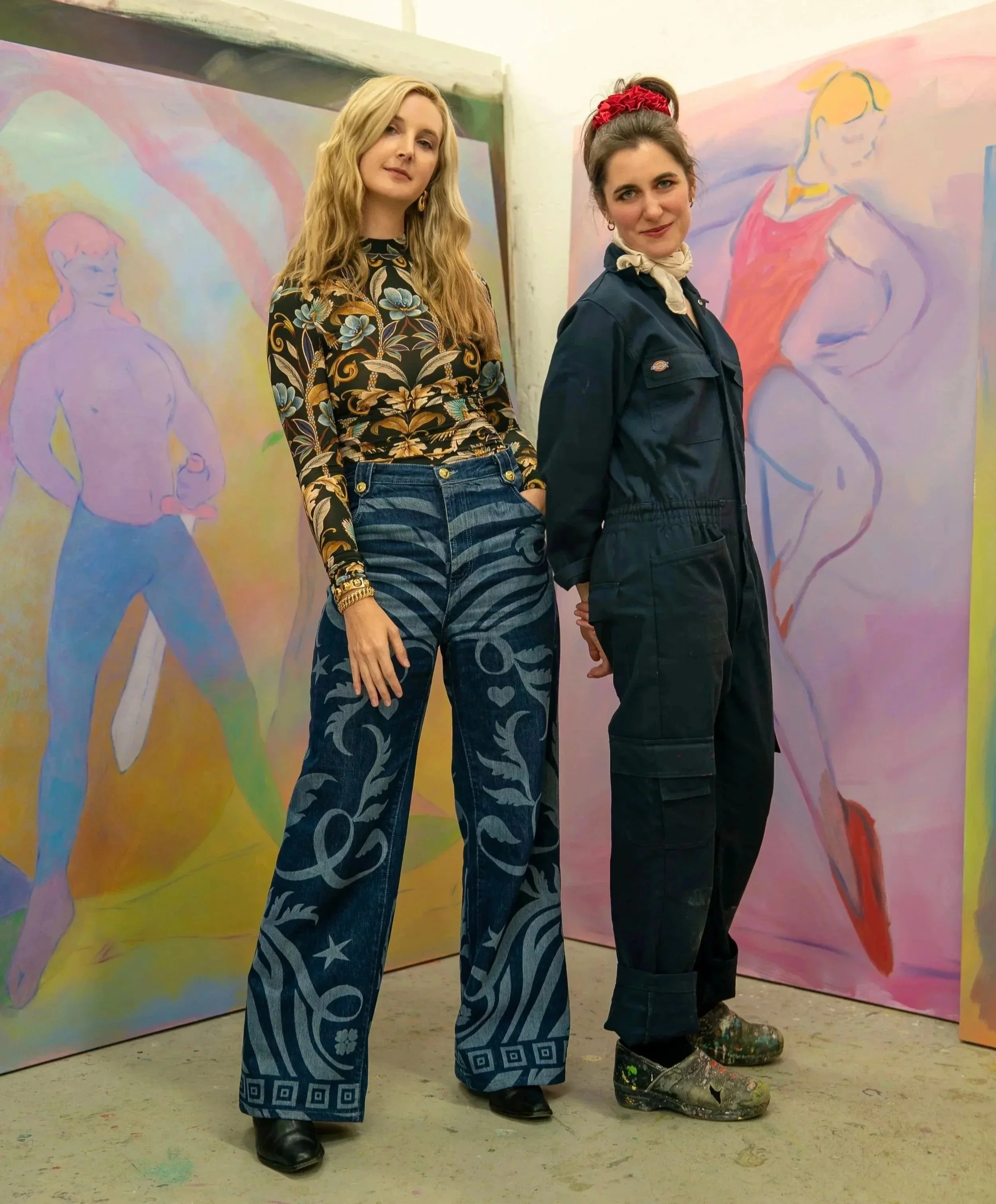 Two women standing in front of abstract pastel-colored paintings. The woman on the left has long blonde hair, is wearing a floral patterned top and wide-leg jeans with star and swirl designs. The woman on the right has dark hair tied back with a red scrunchie, is wearing a dark jumpsuit and colorful patterned boots.