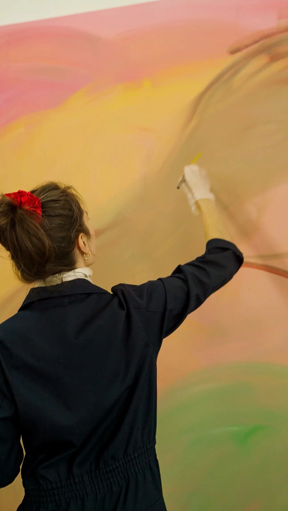 A woman with a red hair scrunchie in her brown hair, wearing a black jacket, is painting on a colorful wall with pastel shades of pink, yellow, and green.