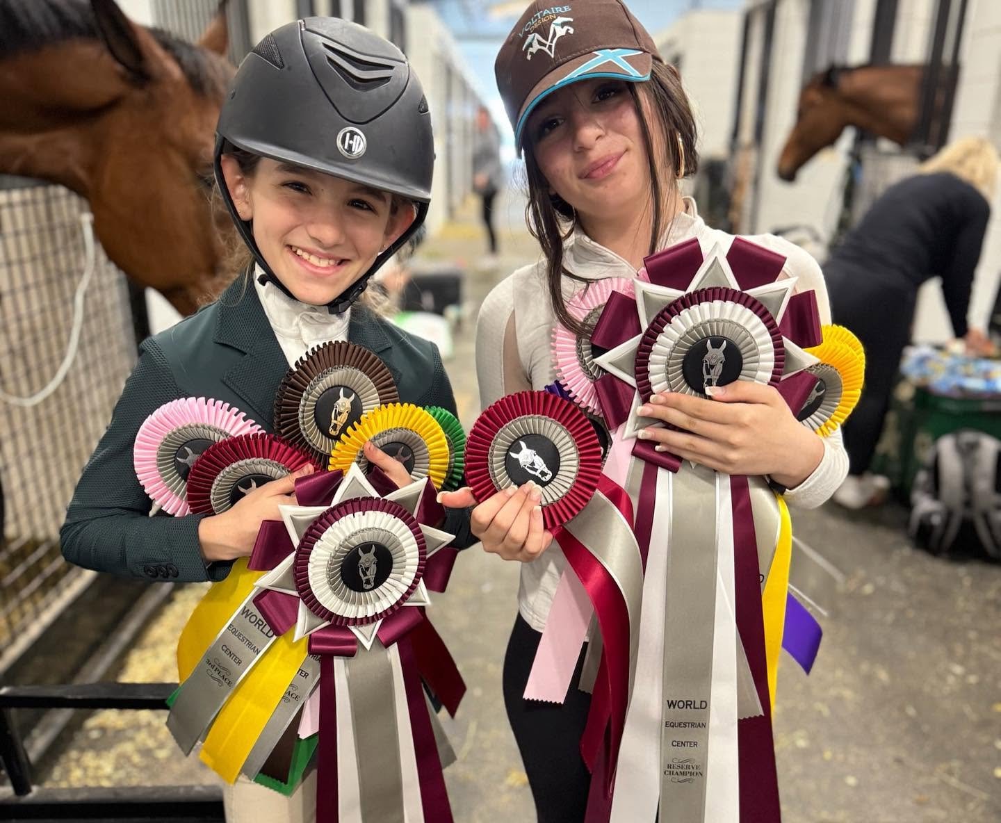 Two young girls, dressed in equestrian attire, smiling and holding numerous colorful ribbons. One girl wears a riding helmet and the other a riding cap. They are in a stable or equestrian center, with horses and other people visible in the background.