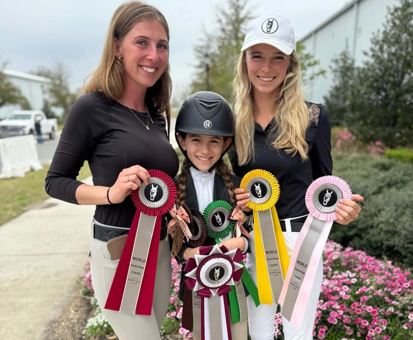 Three females and a young girl standing outdoors, holding colorful award ribbons. The girl is wearing a helmet and riding attire, and the women are dressed in equestrian clothing. Pink flowers and bushes are visible in the background.