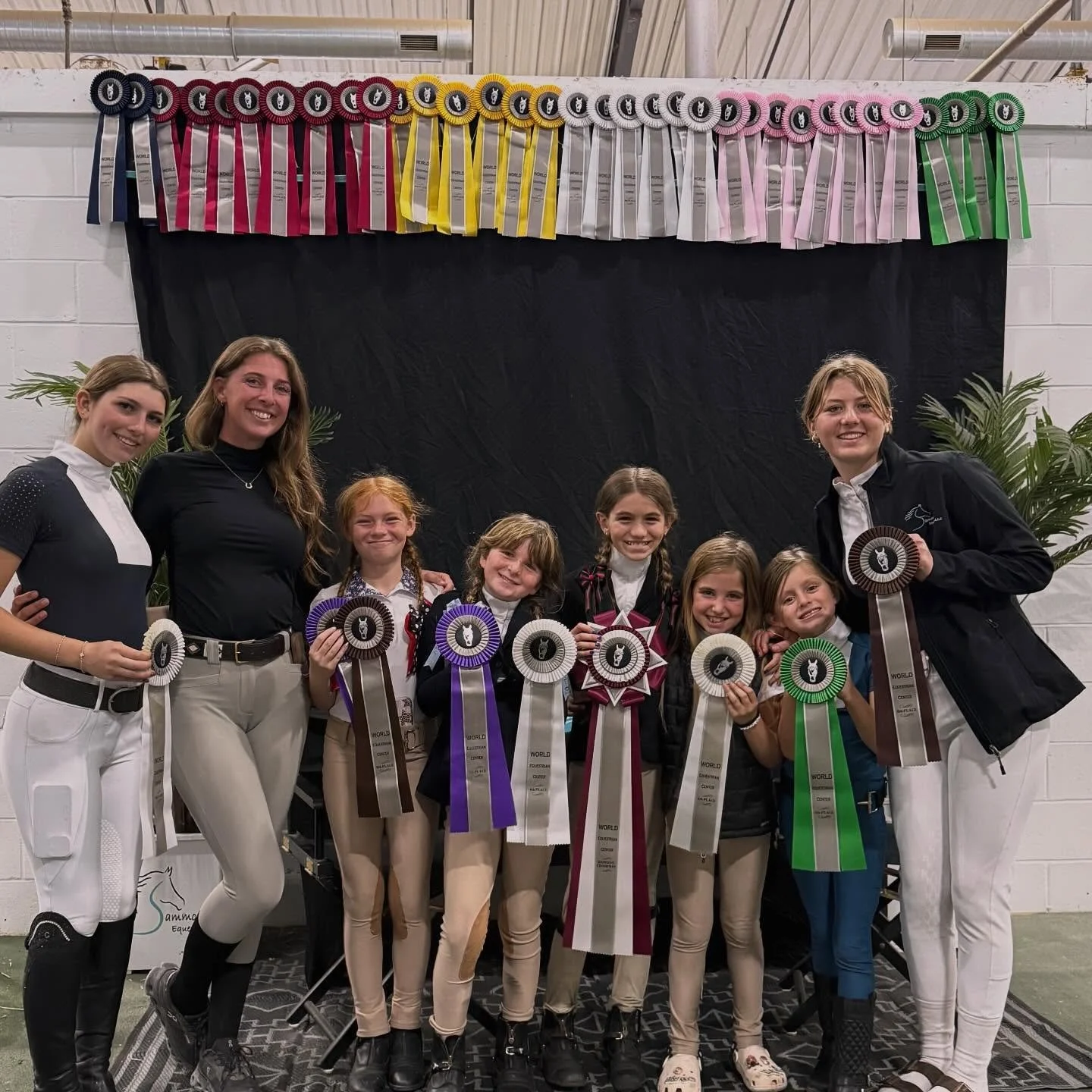 Group of young equestrians and coaches posing with awards at a riding competition, standing in front of a black backdrop with colorful ribbons displayed above and holding rosettes.