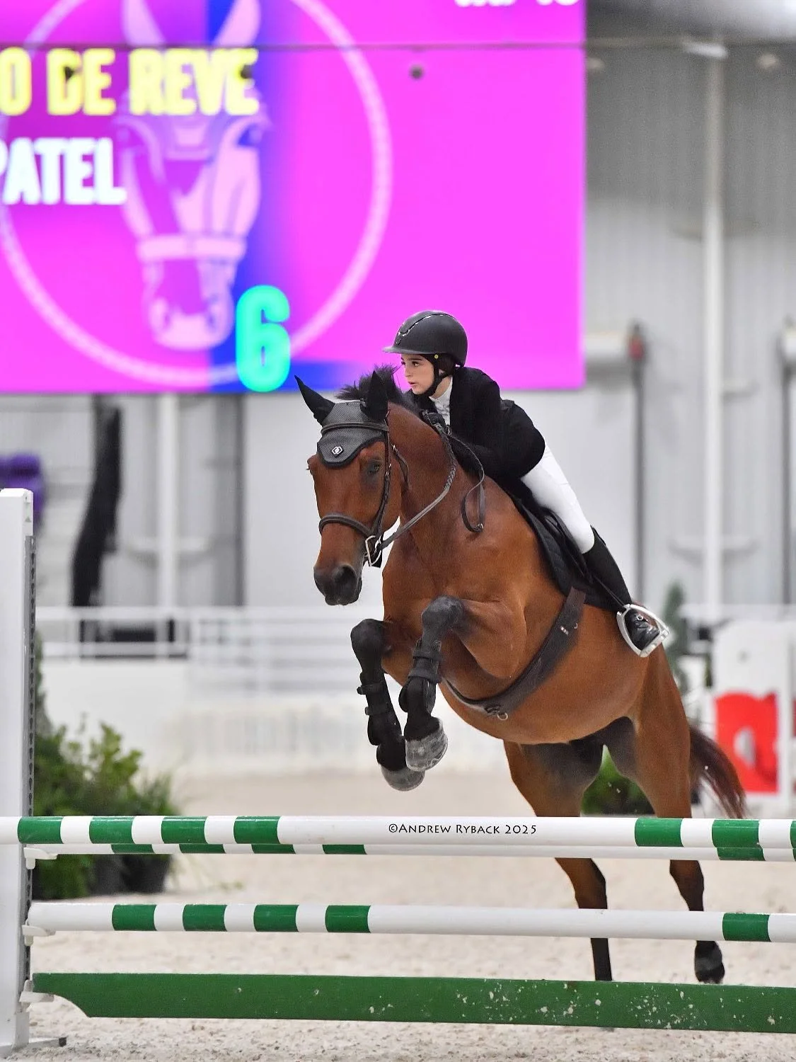 A female equestrian rider dressed in black and white riding attire on a brown horse, jumping over a green and white obstacle at an indoor arena with a pink digital display in the background.