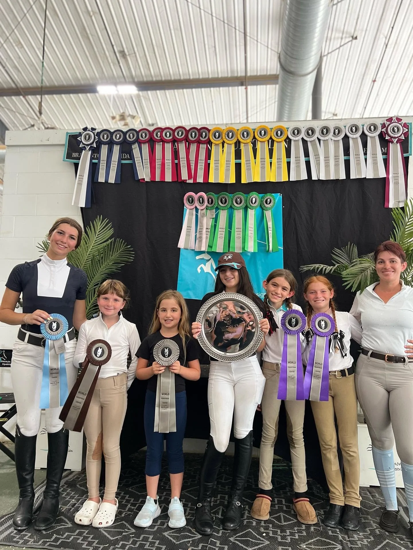 Group of young girls and two women standing indoors, holding blue, brown, black, white, purple, and other colored ribbons in front of a black backdrop with ribbons and plants on either side. They are dressed in equestrian attire and smiling after a competition.