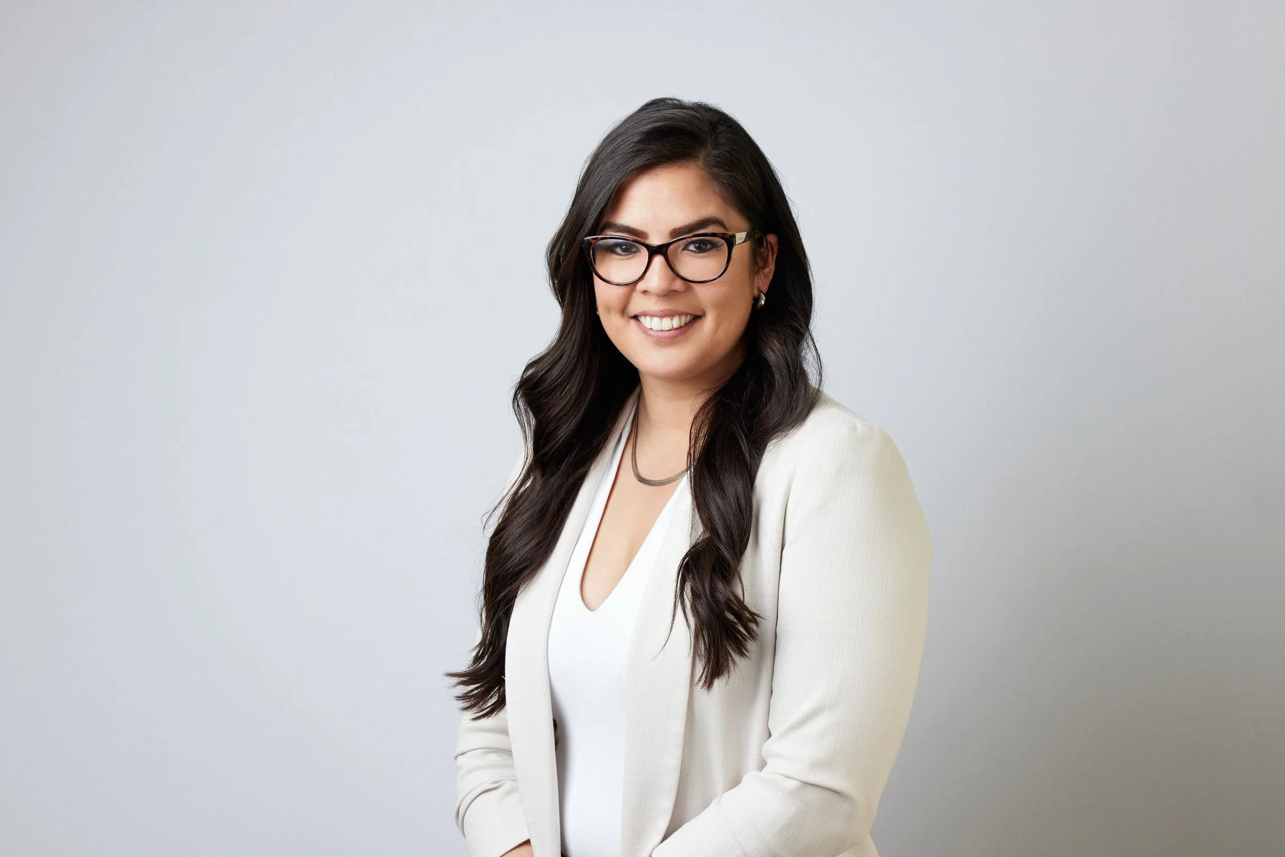 A professional woman with long dark hair, glasses, and a white blazer smiling against a plain white background.