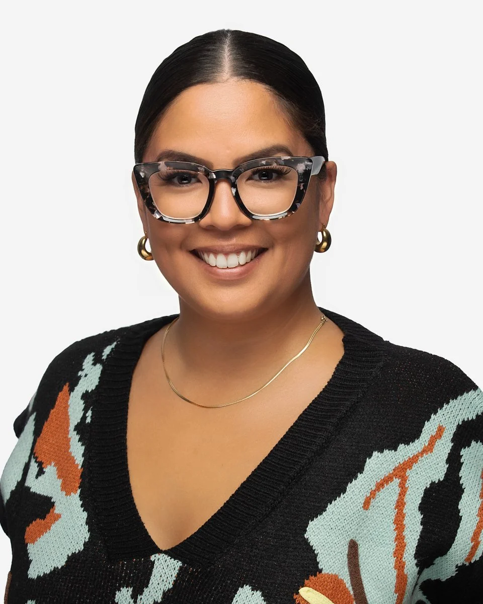 A woman with long, wavy dark hair wearing glasses, a white blazer, and a white top, smiling at the camera against a plain white background.