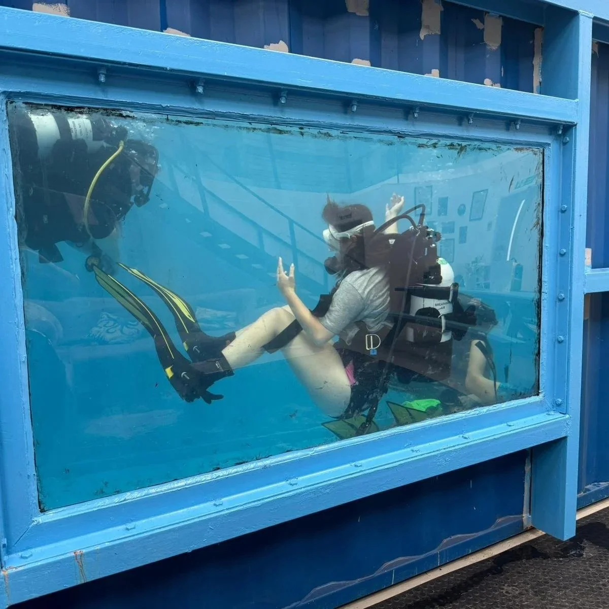 A person scuba diving inside a glass tank, giving a peace sign with one hand, with scuba gear and fins, outdoors.