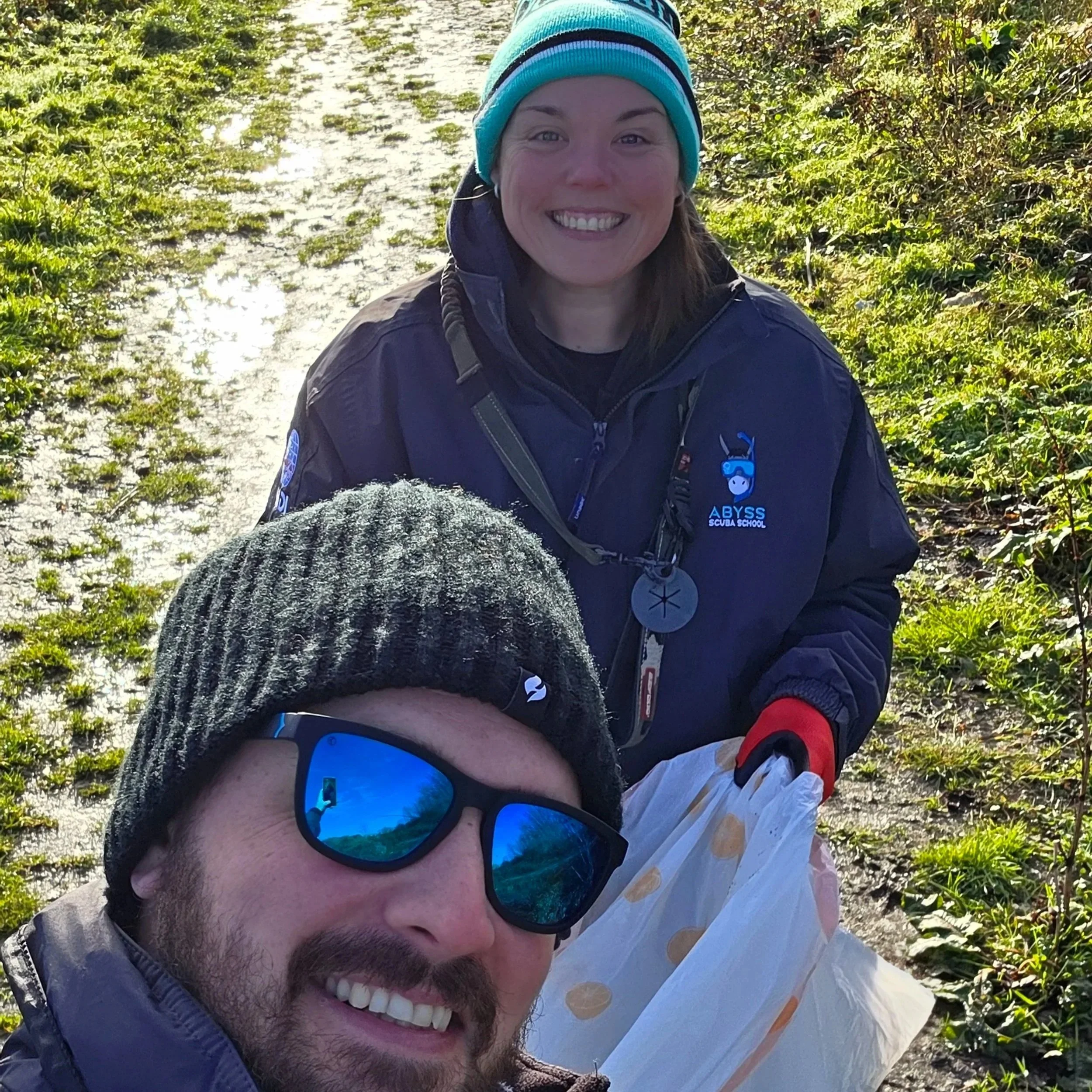 A man and a woman taking a selfie outdoors. The woman is smiling, wearing a blue beanie and a navy jacket with a diving school logo, and standing behind the man. The man is wearing sunglasses, a black beanie, and a jacket, and is in the foreground.