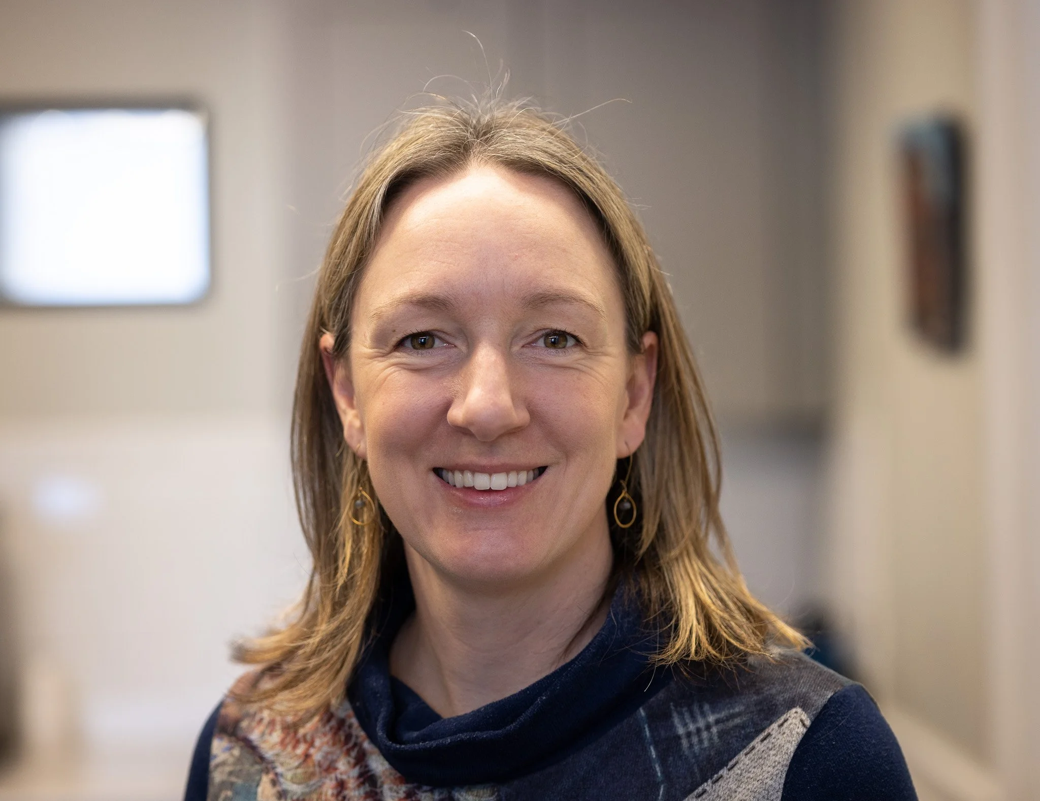 Smiling woman with shoulder-length brown hair wearing earrings and a patterned scarf in an indoor setting.