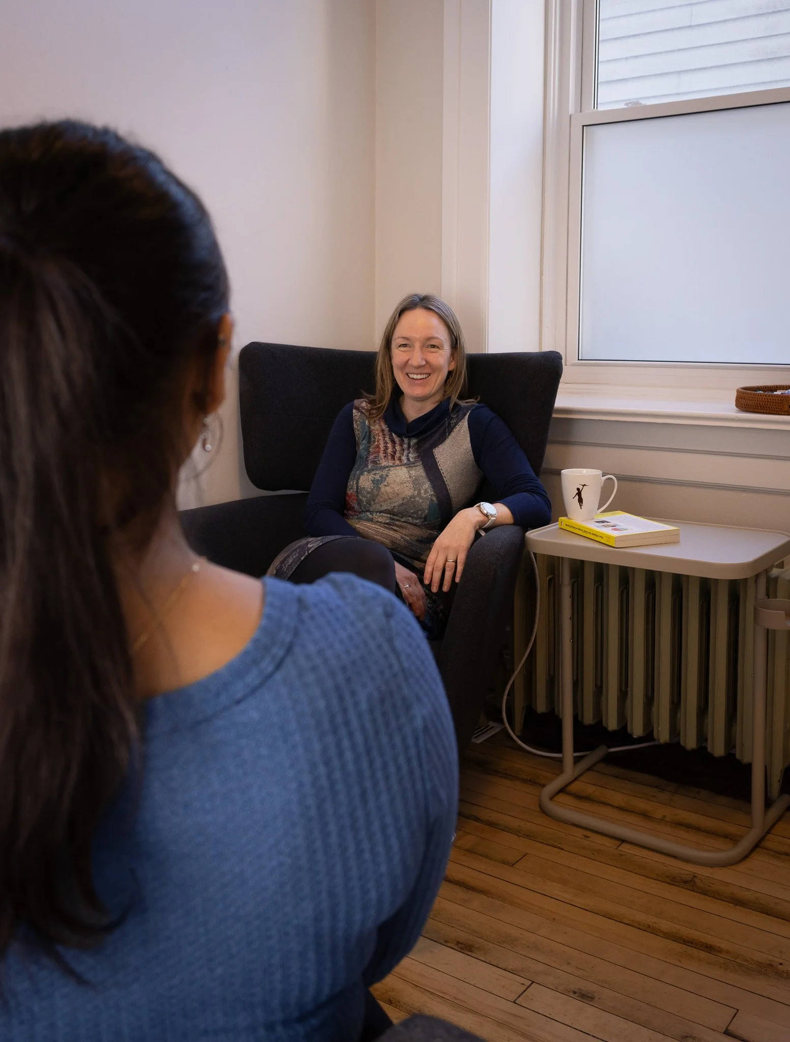 Two women having a conversation in a room with wooden floors, a black armchair, and a small table near a window. One woman is sitting in the armchair, smiling, while the other woman is partially visible from behind.