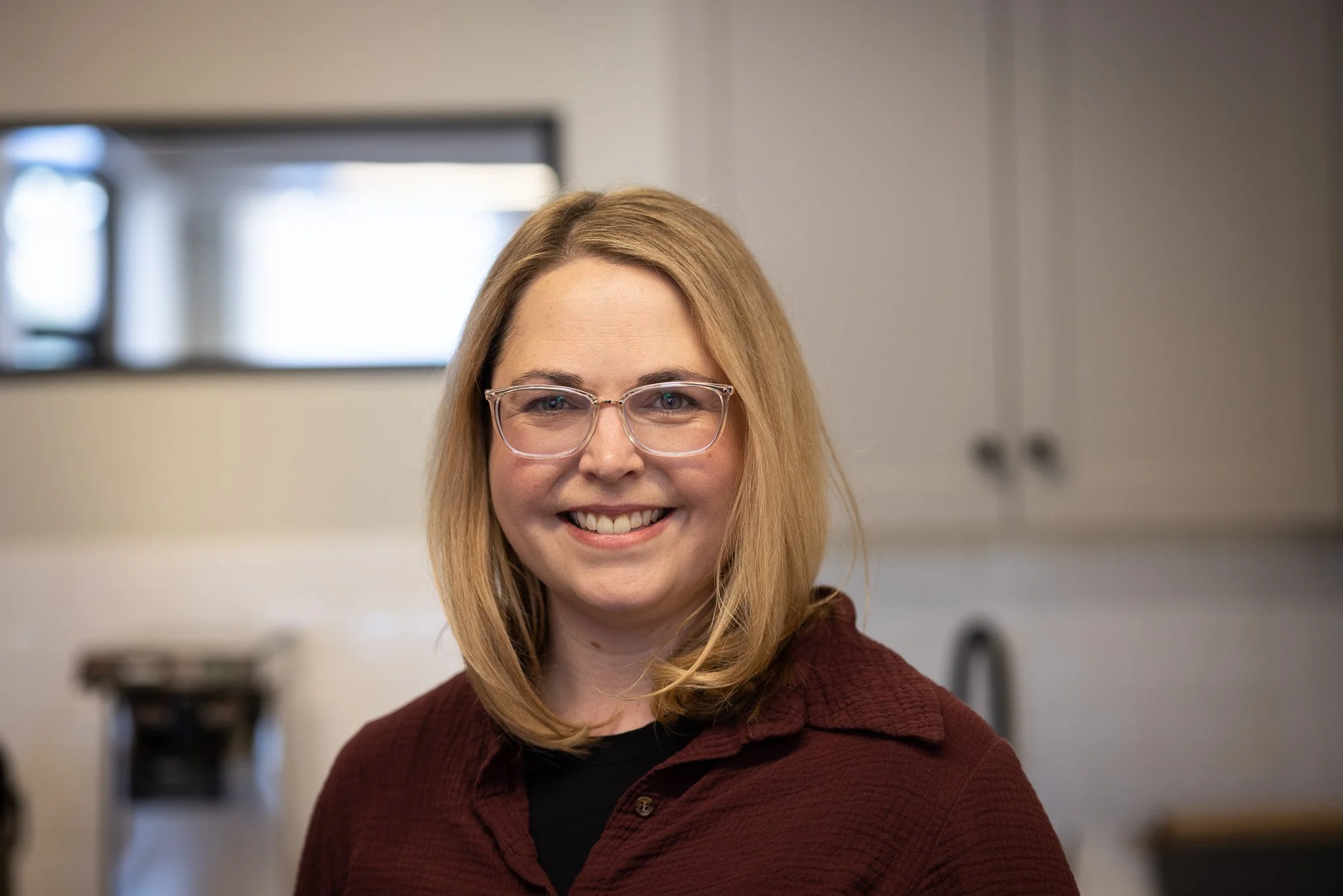 A woman with shoulder-length blonde hair, wearing glasses and a maroon jacket, smiling indoors.