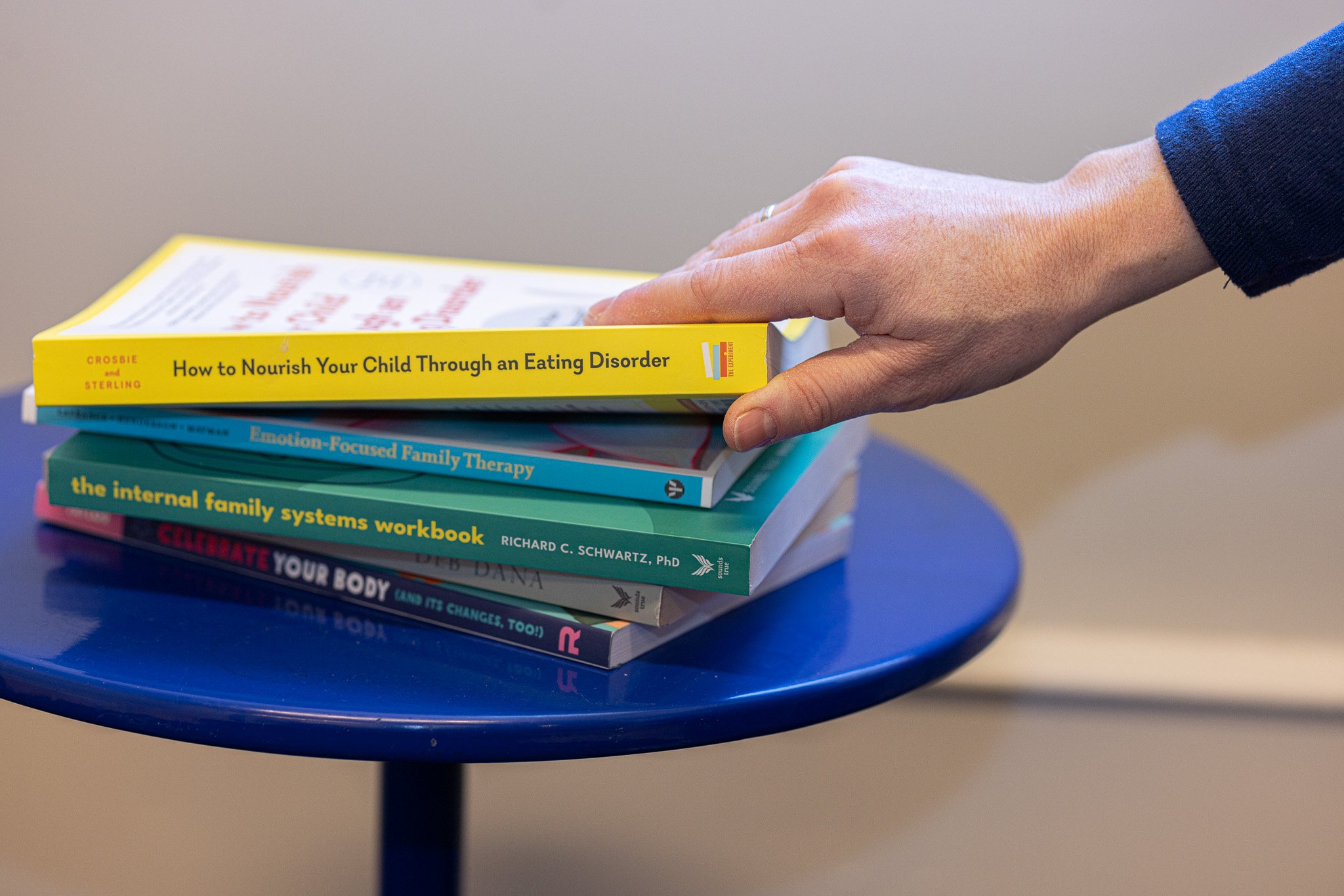 A hand with a blue sleeve resting on top of a stack of five books on a small blue table. The books are about mental health, family therapy, and eating disorders.