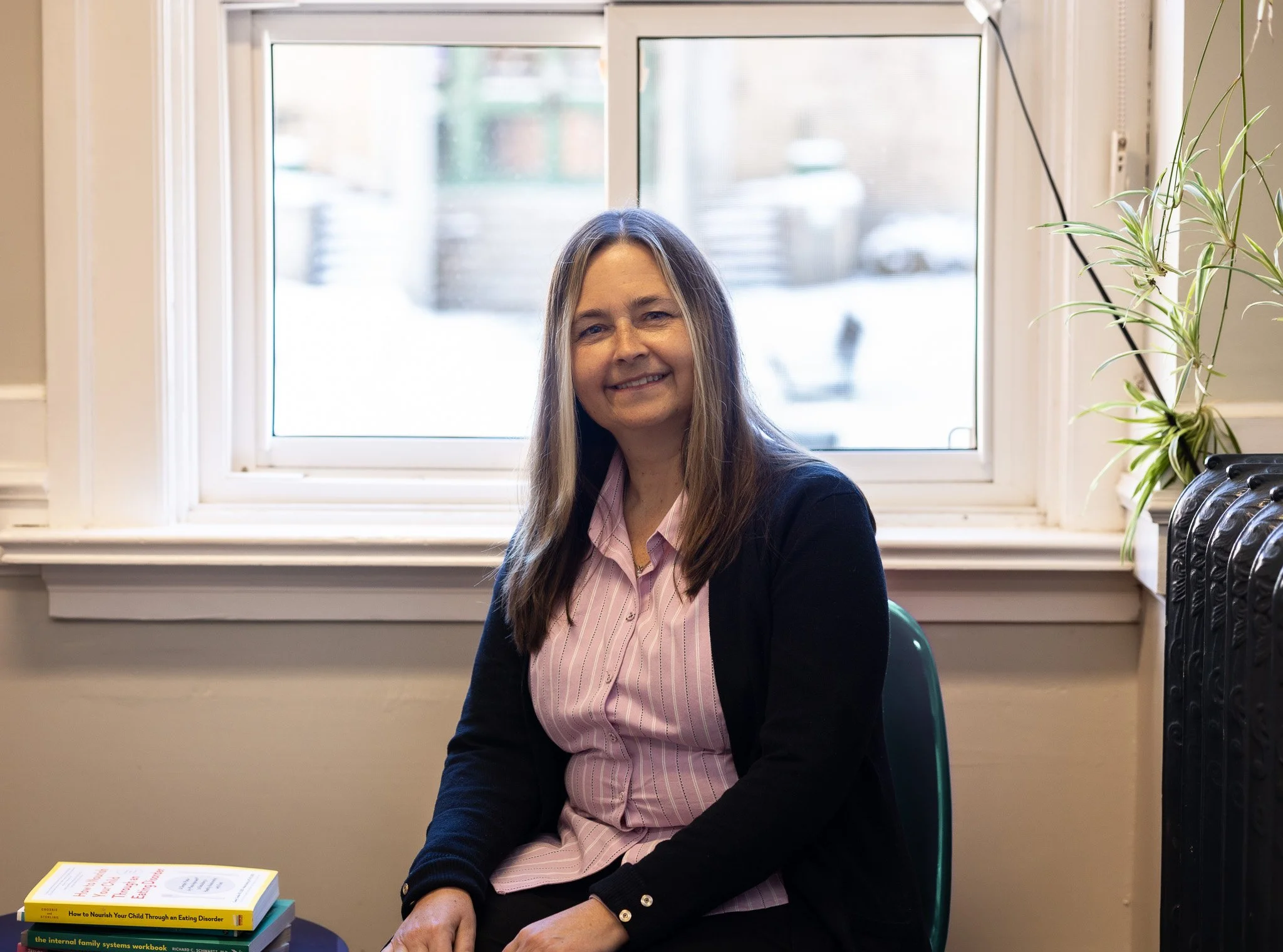 A woman with long brown hair smiling, sitting in a room with a large window behind her and a stack of books on a table in front of her.