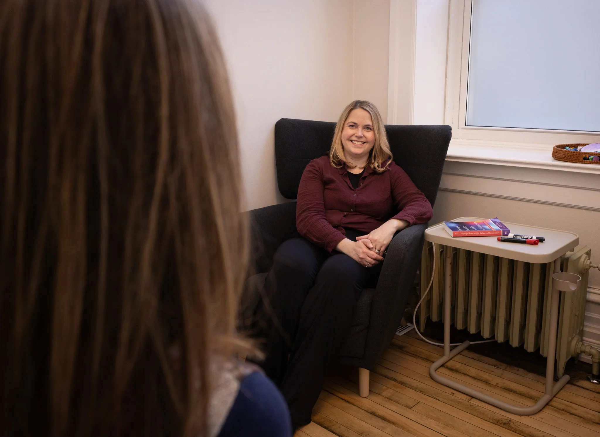 A woman with shoulder-length blonde hair sitting in a black armchair, smiling during an interview or conversation in a room with a window and a radiator, with a side table holding books and markers nearby.