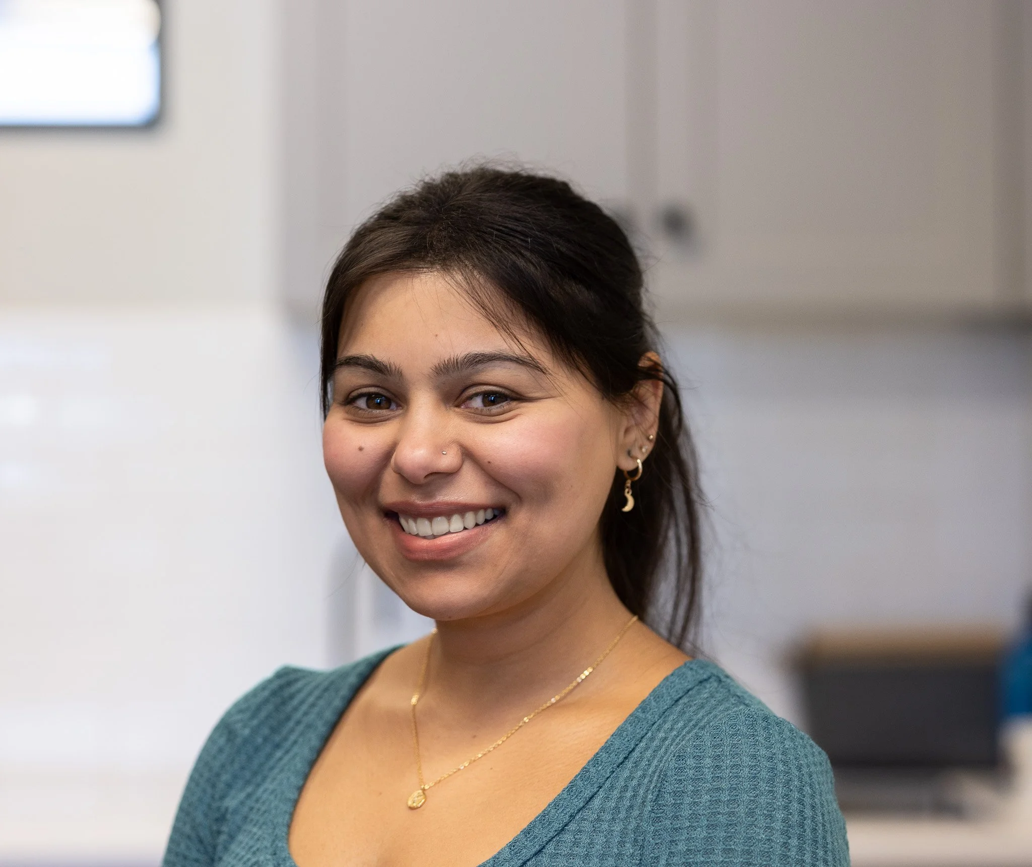 A smiling woman with dark hair pulled back, wearing a teal top, gold jewelry, and earrings, in an indoor setting.