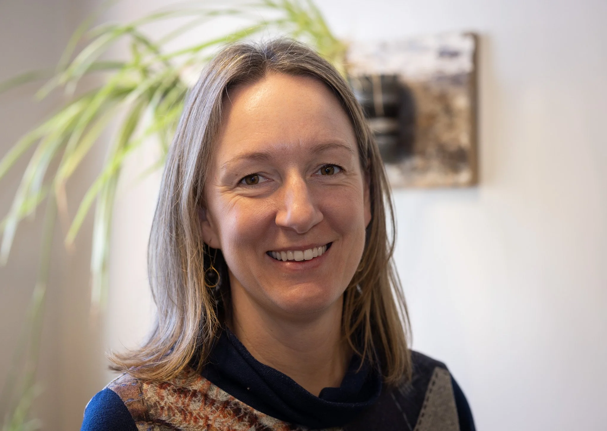 A woman with shoulder-length brown hair smiling indoors with a plant and abstract art on the wall behind her.