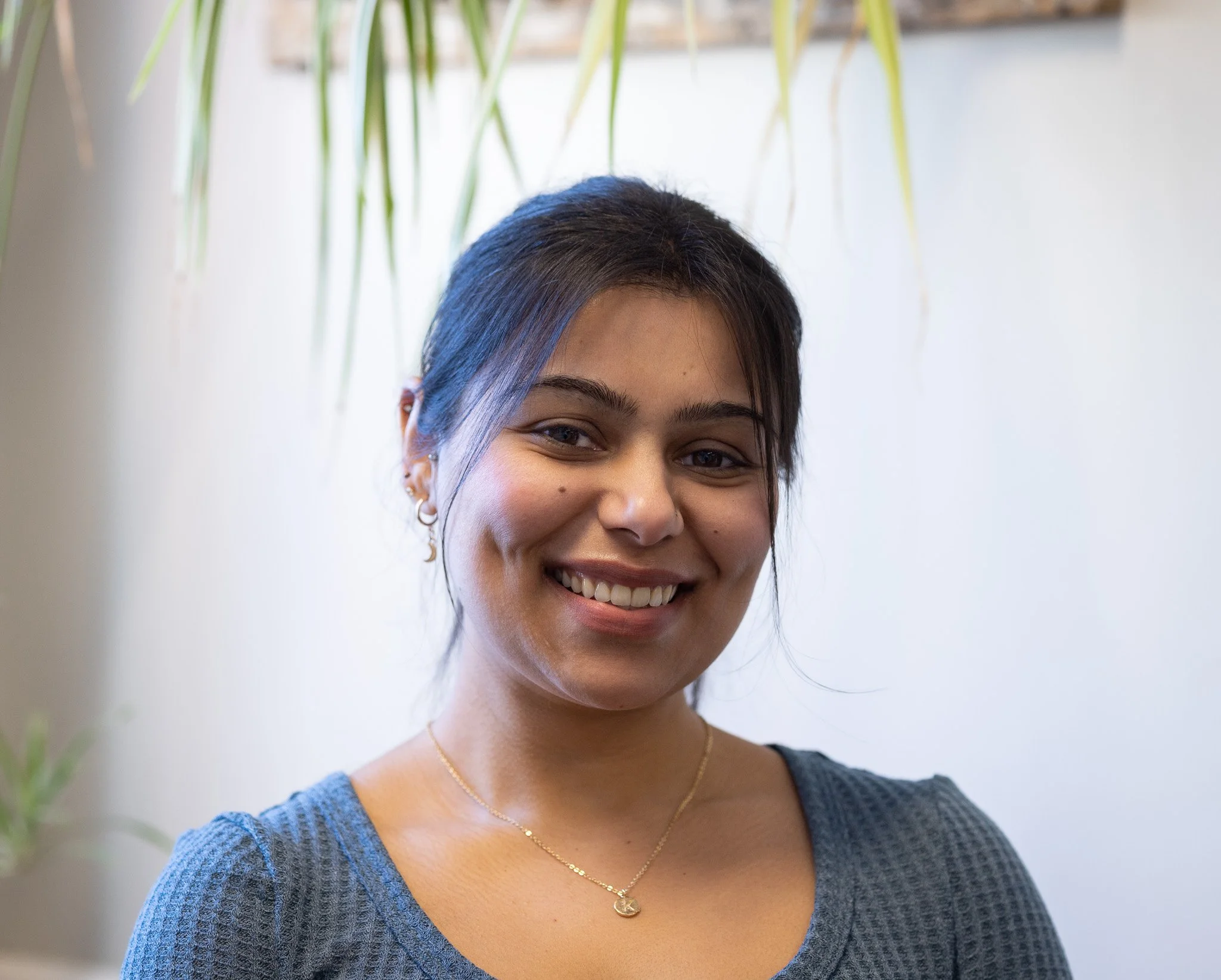 A woman with dark hair, smiling, wearing a gray textured top, gold jewelry, standing indoors with a plant in the background.