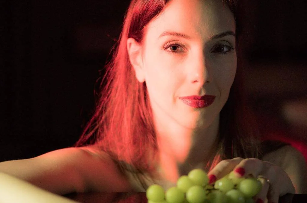 Mulher com cabelo vermelho e lábios vermelhos, segurando uvas verdes, com iluminação suave.