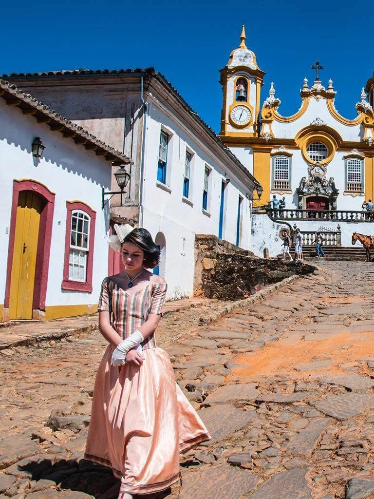 Fotografia fine art de mulher com vestido colonial caminhando por rua de pedra em Tiradentes, Minas Gerais — obra de Alexandre Horta e Silva que une história, identidade e estética brasileira.