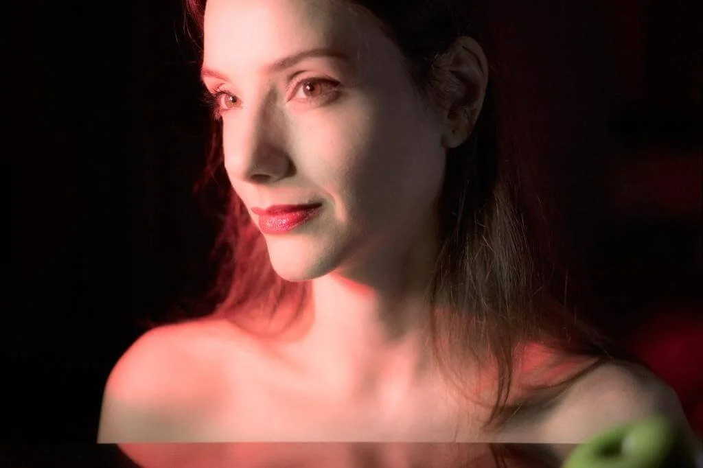 Retrato de uma mulher com pele clara, cabelo castanho e expressão suave, com iluminação suave e fundo escuro.