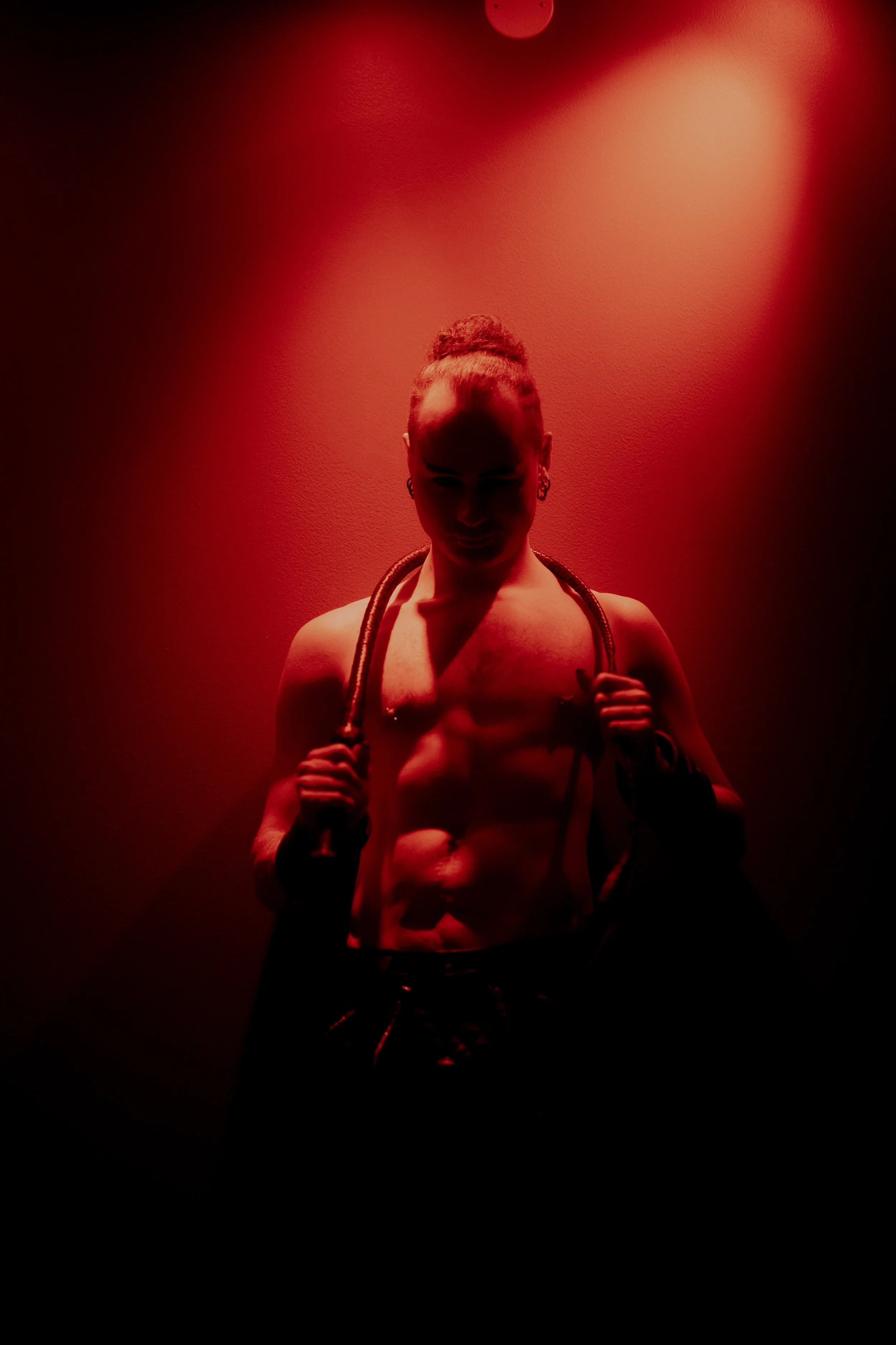 A shirtless muscular Dominant holding a leather whip over their shoulders, illuminated by red lighting, with a shadowed face and a dark background. - Picture by Iromie