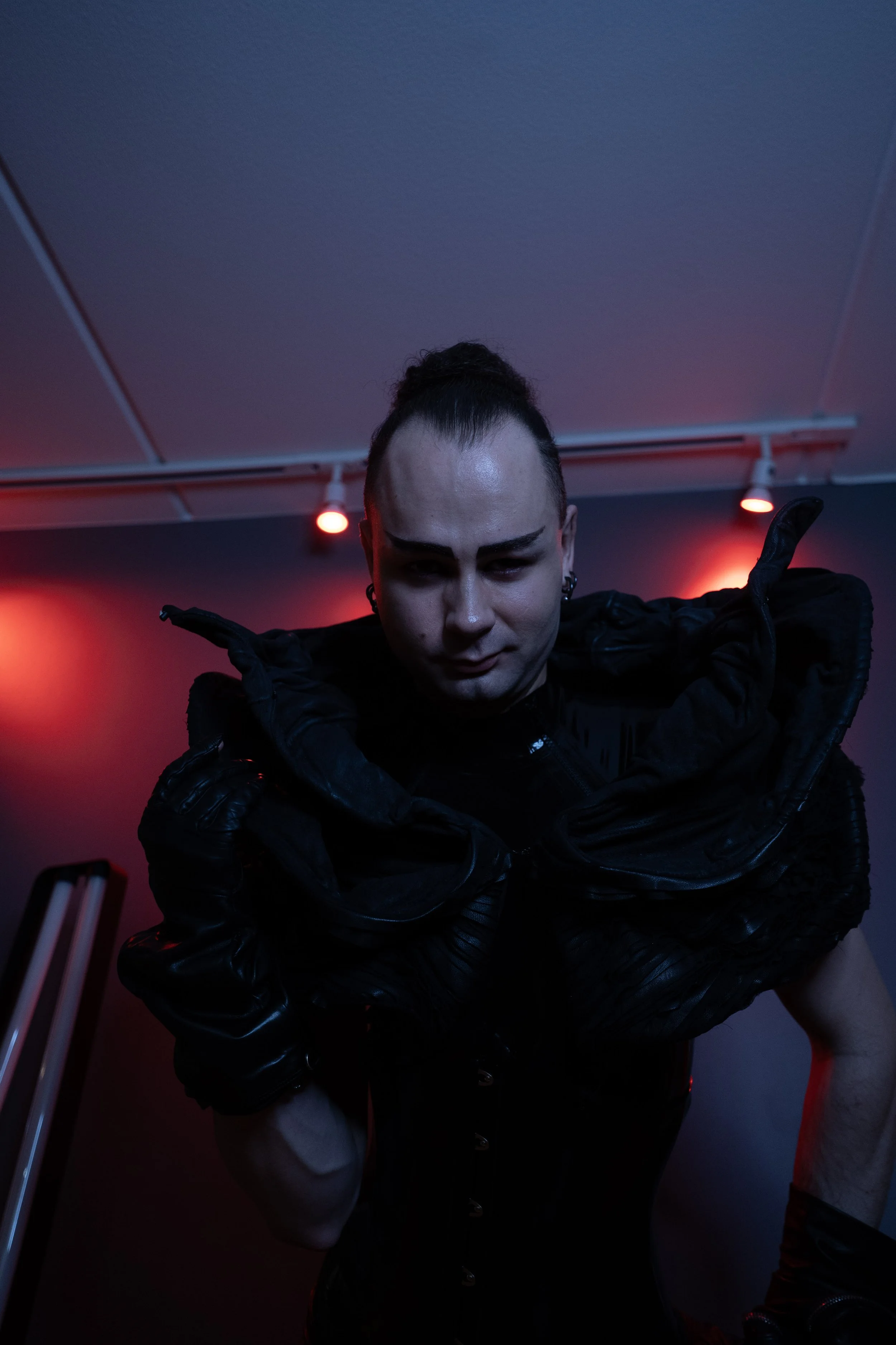 Dominatrix with dark makeup and styled hair, wearing a black, metallic, futuristic-looking latex outfit posing against a red gradient background. -Picture taken by Iromie