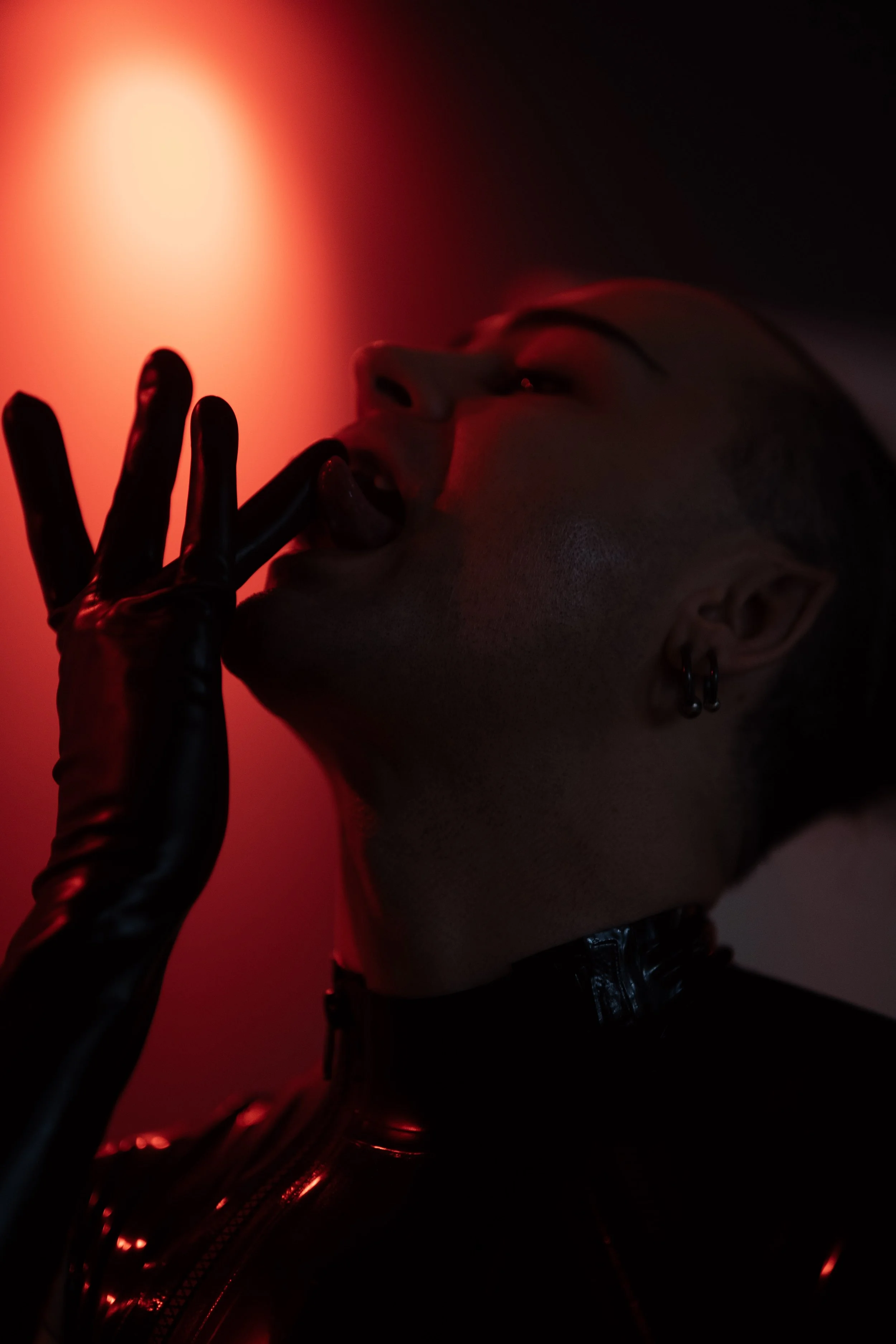 A person wearing a black latex gloves, illuminated by red light, with their head tilted back and lips slightly parted revealing a split tongue. -Picture taken by Iromie