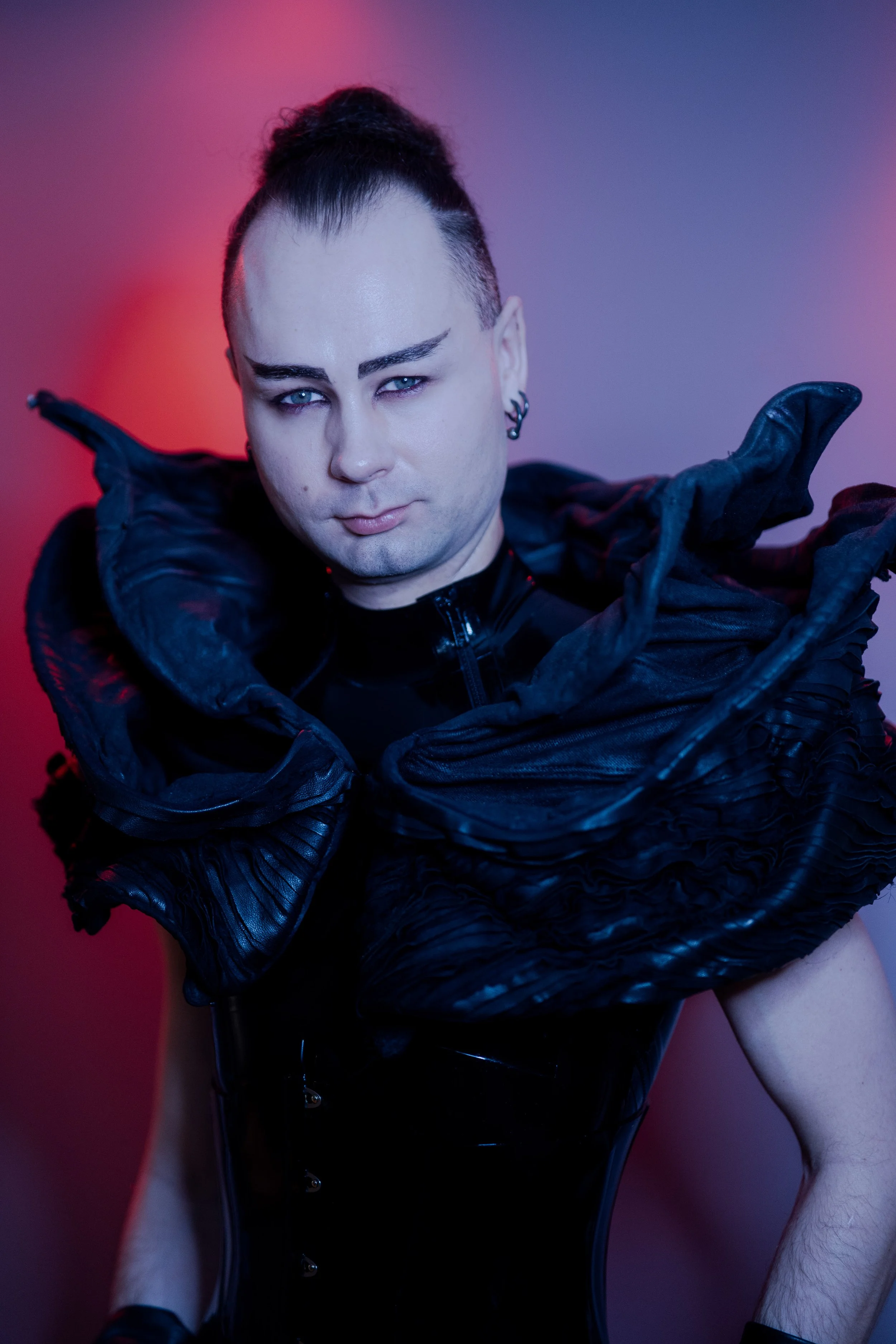 Dominatrix with dark makeup and styled hair, wearing a black, metallic, futuristic-looking latex outfit posing against a red gradient background. -Picture taken by Iromie
