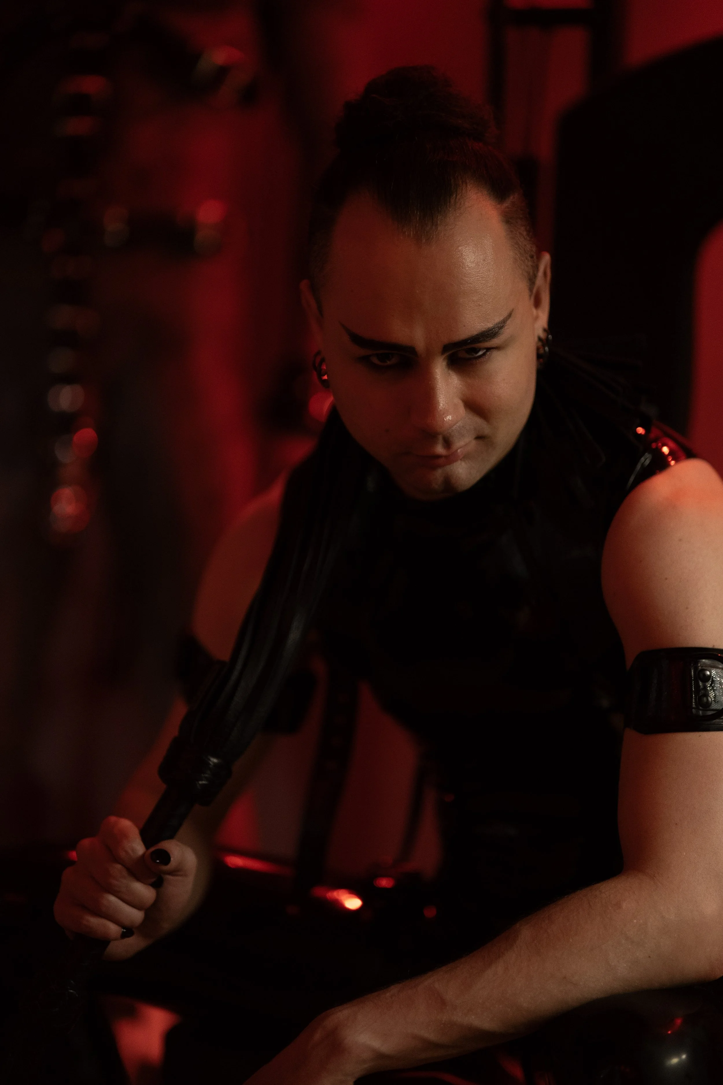 Professional Dominant (male escort) wearing a sleevelees latex catsuit and holding a leather flogger. Picture taken by Iromie