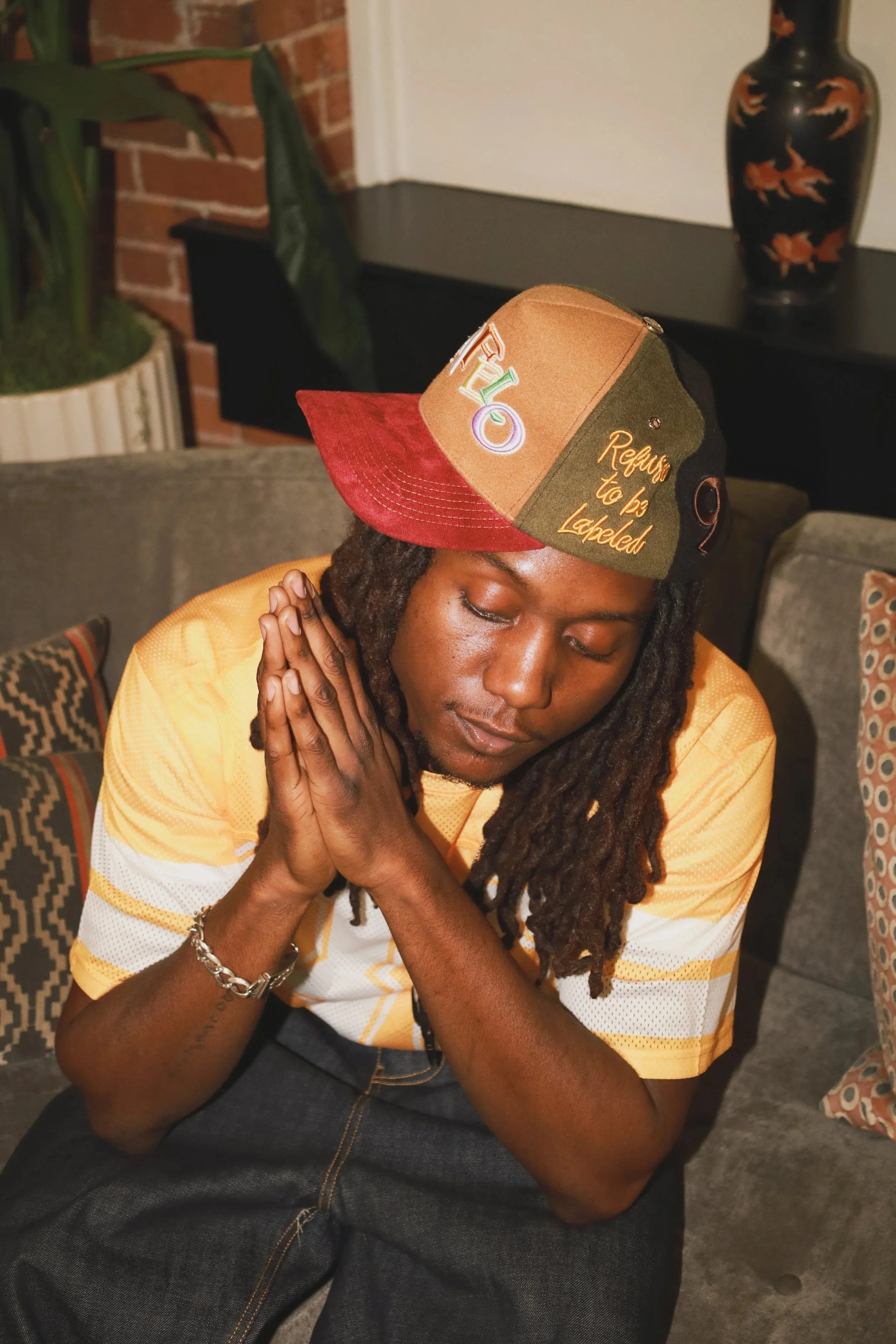 A young man with dreadlocks sitting on a couch with his eyes closed and hands pressed together in a prayer position, wearing a colorful baseball cap and a yellow sports jersey.