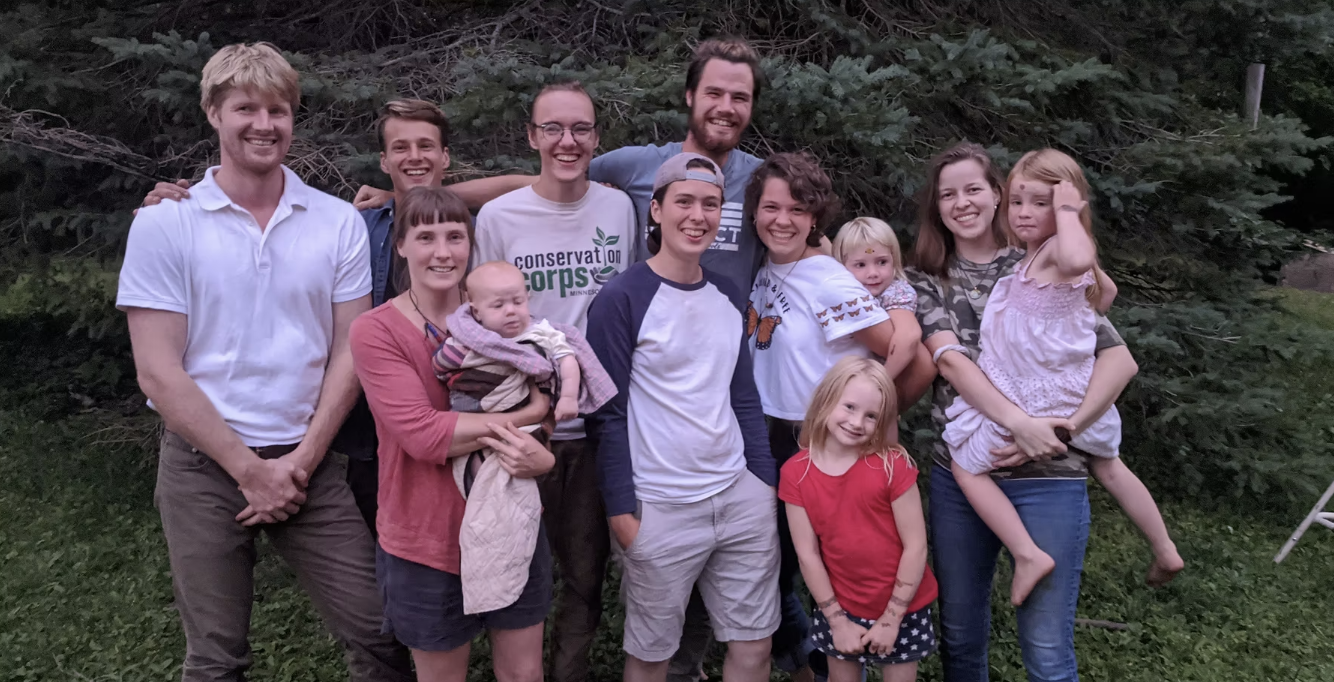 Six people stand outdoors in front of greenery, posing for a photo with a large fluffy dog in the foreground.