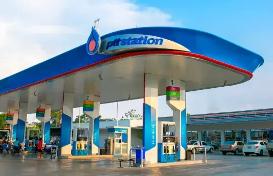 A gas station with a Blue and white canopy displaying the PTT station logo, multiple fuel pumps, and several cars refueling under a partly cloudy sky.